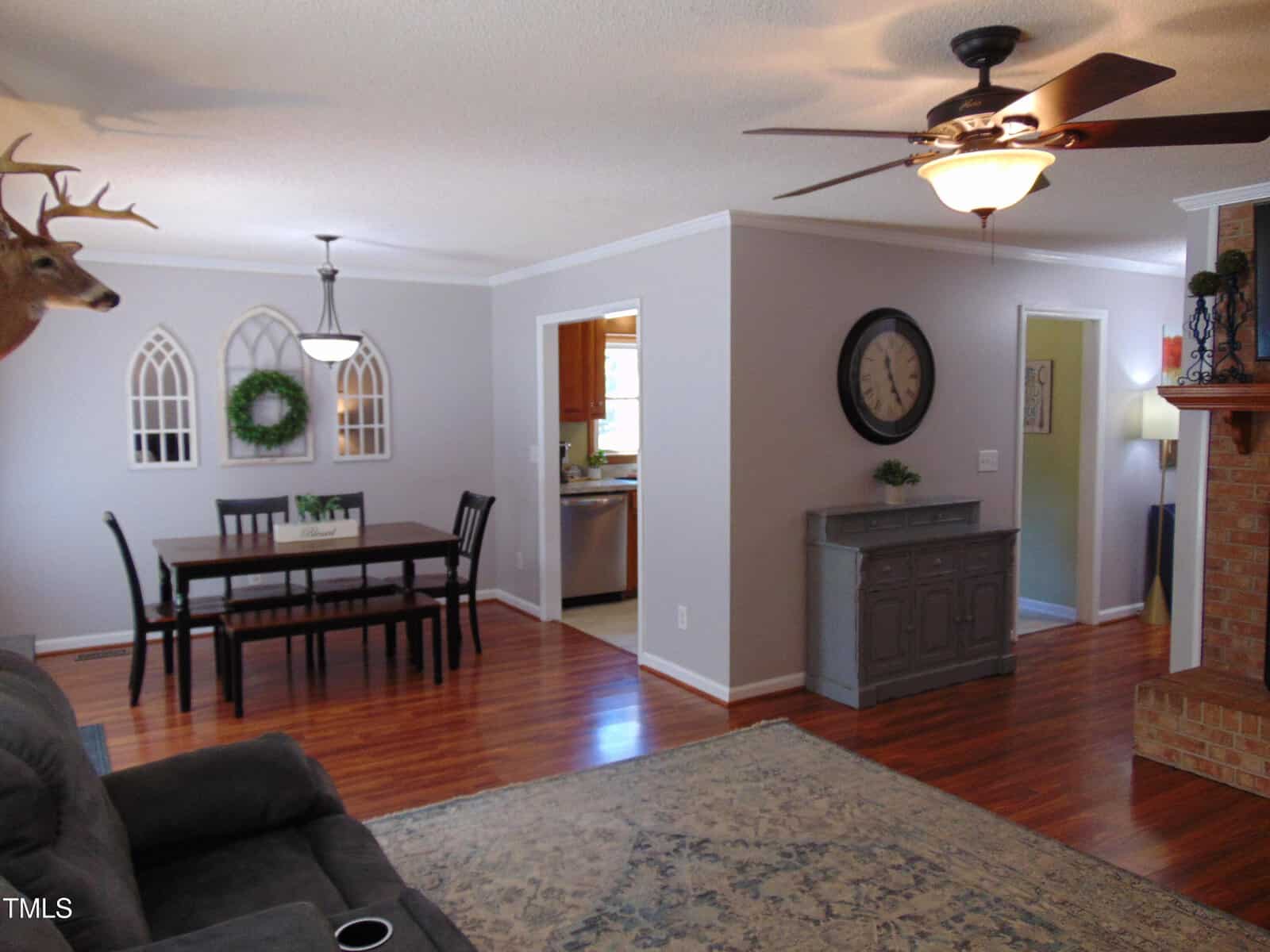 A living room with wood floors, a brick fireplace, ceiling fan, and gray sofa. Adjacent dining area has a table with four chairs and decorative wall mirrors. A gray sideboard with a clock is near the entryway to the kitchen.