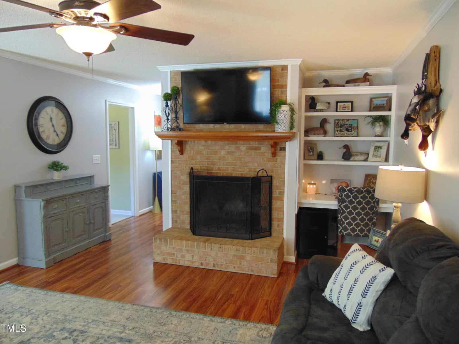 A cozy living room with a brick fireplace, TV above the mantel, shelves with decor items, a dark couch with patterned pillows, a gray sideboard, a wall clock, and wood flooring. A ceiling fan and lamp provide lighting.