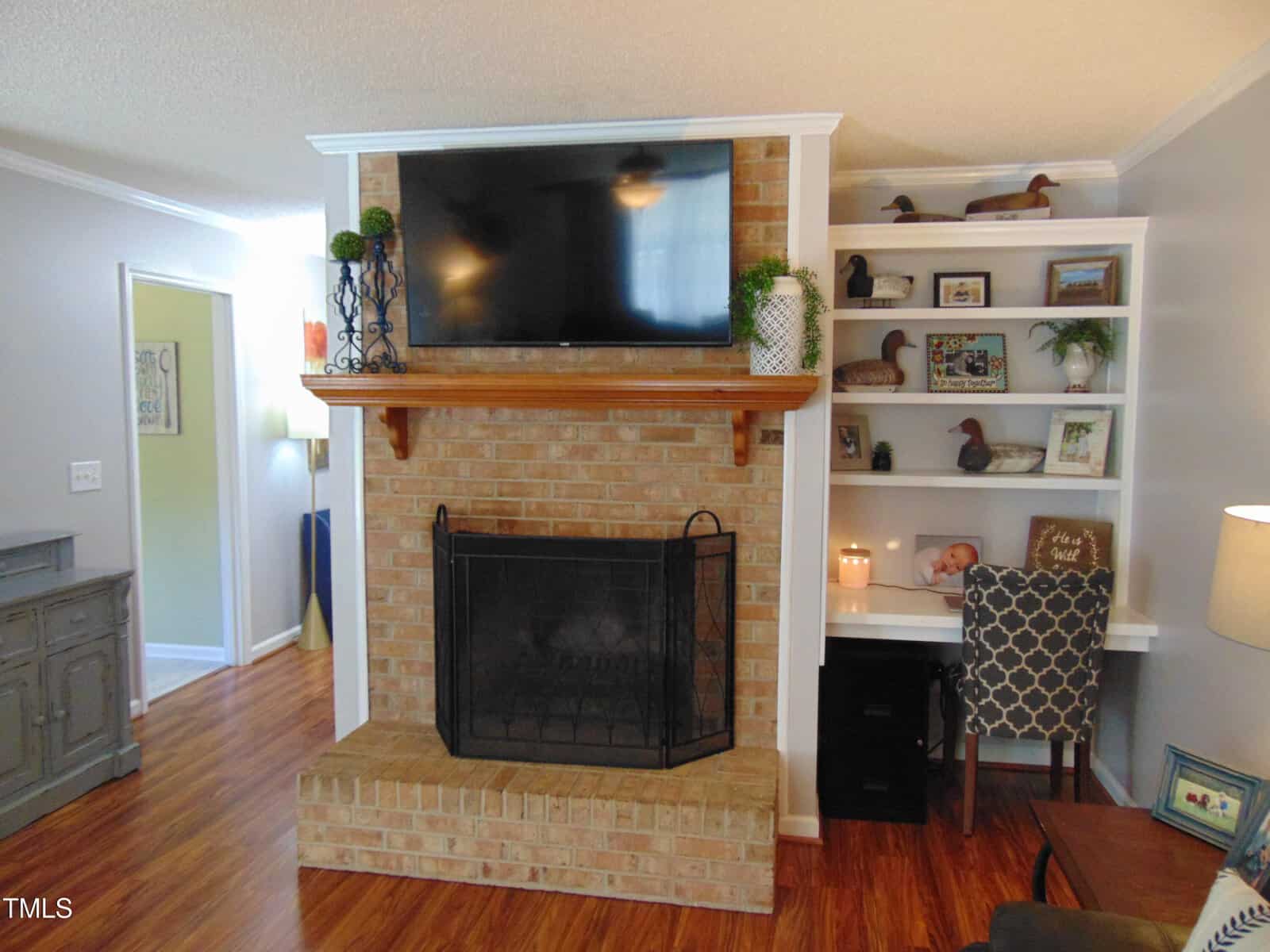 A living room with a brick fireplace, a TV above it, built-in shelves and a small desk to the right, wood flooring, and neutral-colored walls. Decor includes plants, framed photos, and decorative items.