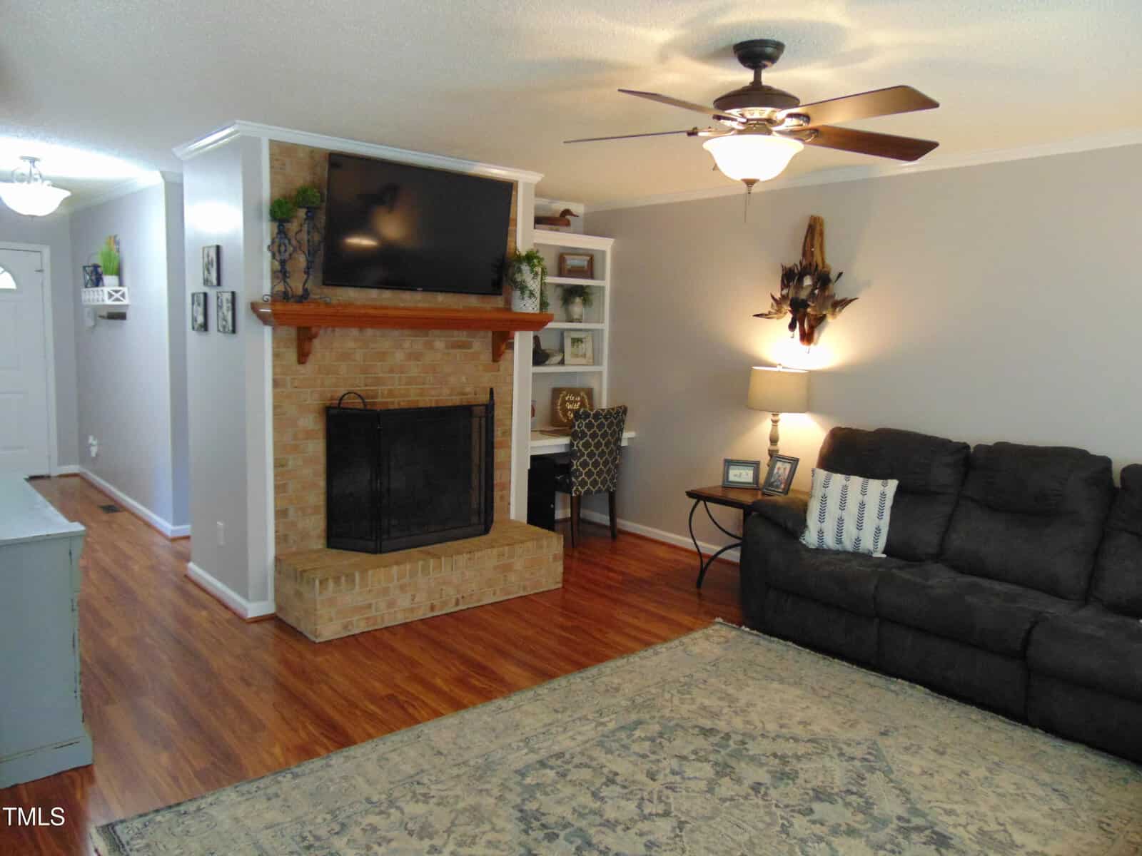 A living room with wood floors, a brick fireplace, a wall-mounted TV, a dark sofa, a ceiling fan, a decorative rug, and a small reading nook with a desk and chair beside the fireplace.