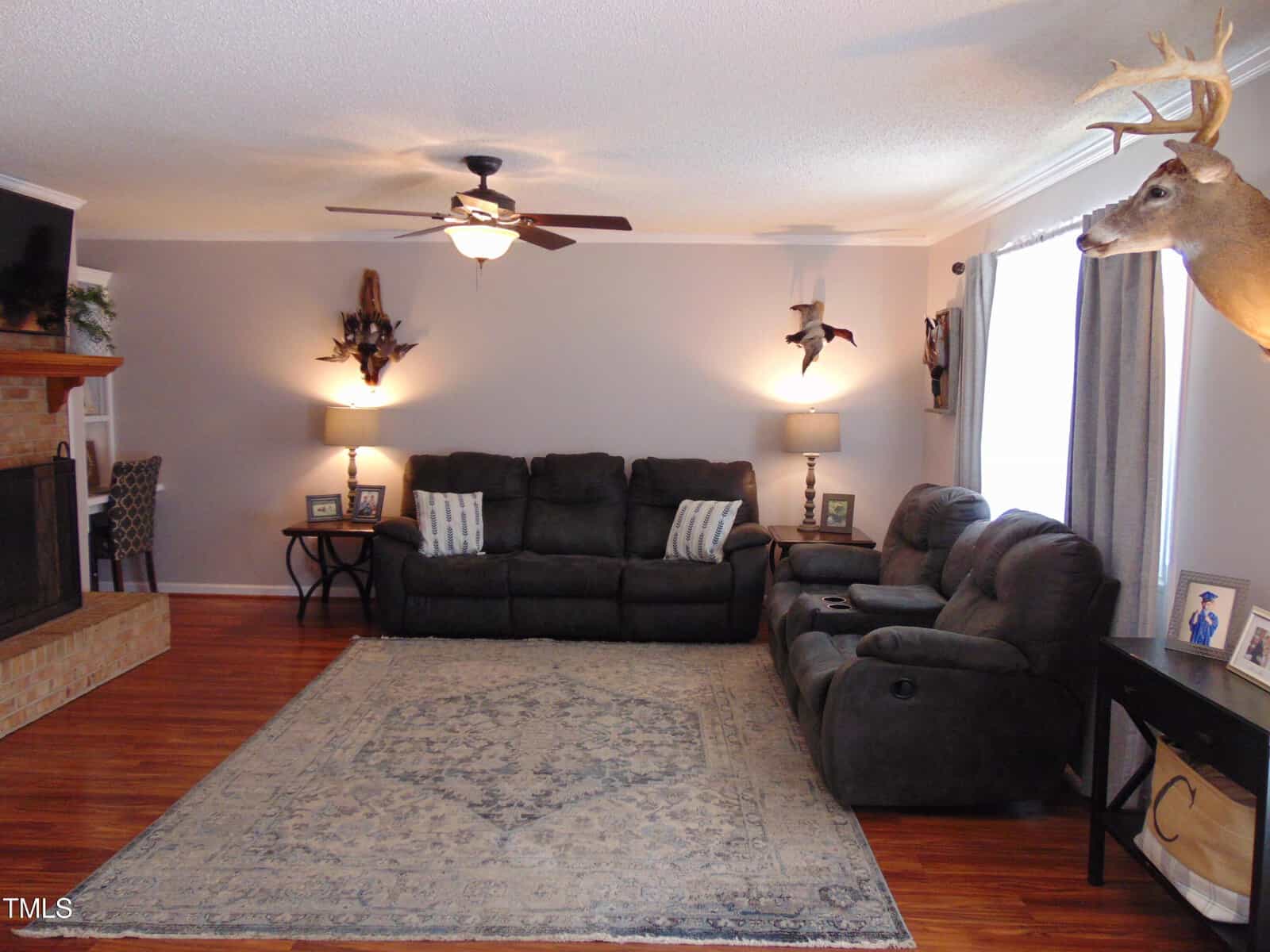 A cozy living room with gray couches, a patterned rug, mounted deer heads, a ceiling fan, lamps, and a brick fireplace. Hardwood floors and gray walls create a warm, inviting atmosphere.