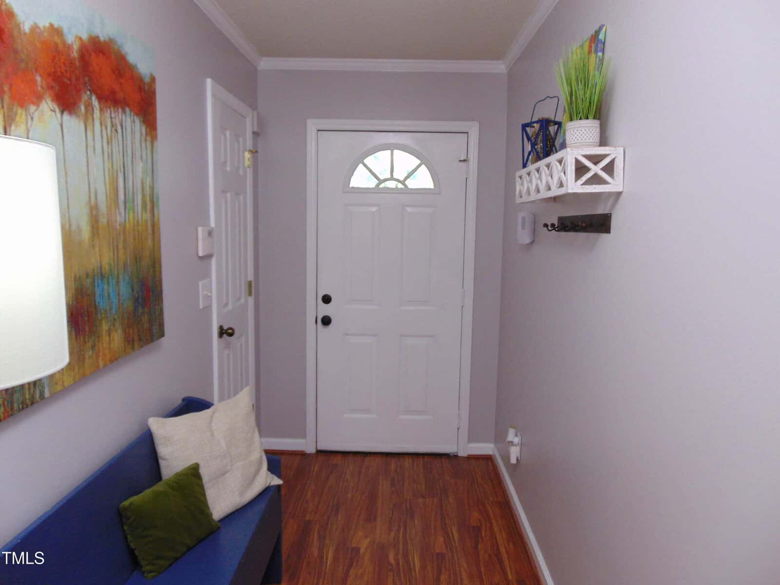 A small entryway with a white front door, colorful abstract painting on the left wall, blue bench with pillows, and a white wall shelf holding decor items on the right. The floor is a medium wood tone.