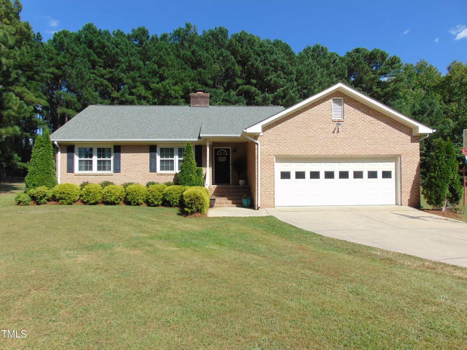 Located at 15 Gina Lee Terrace in Louisburg, this single-story brick house features a gray roof, white trim, two-car garage, and neatly trimmed bushes on a grassy lawn with tall trees and a clear blue sky as the backdrop.