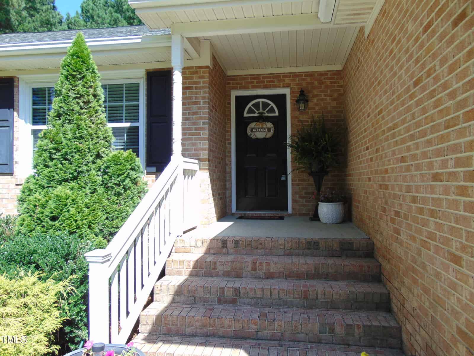 A brick house with a small covered porch, white railing, and steps leading to a black front door with a welcome sign. There are green shrubs and potted plants by the entrance.