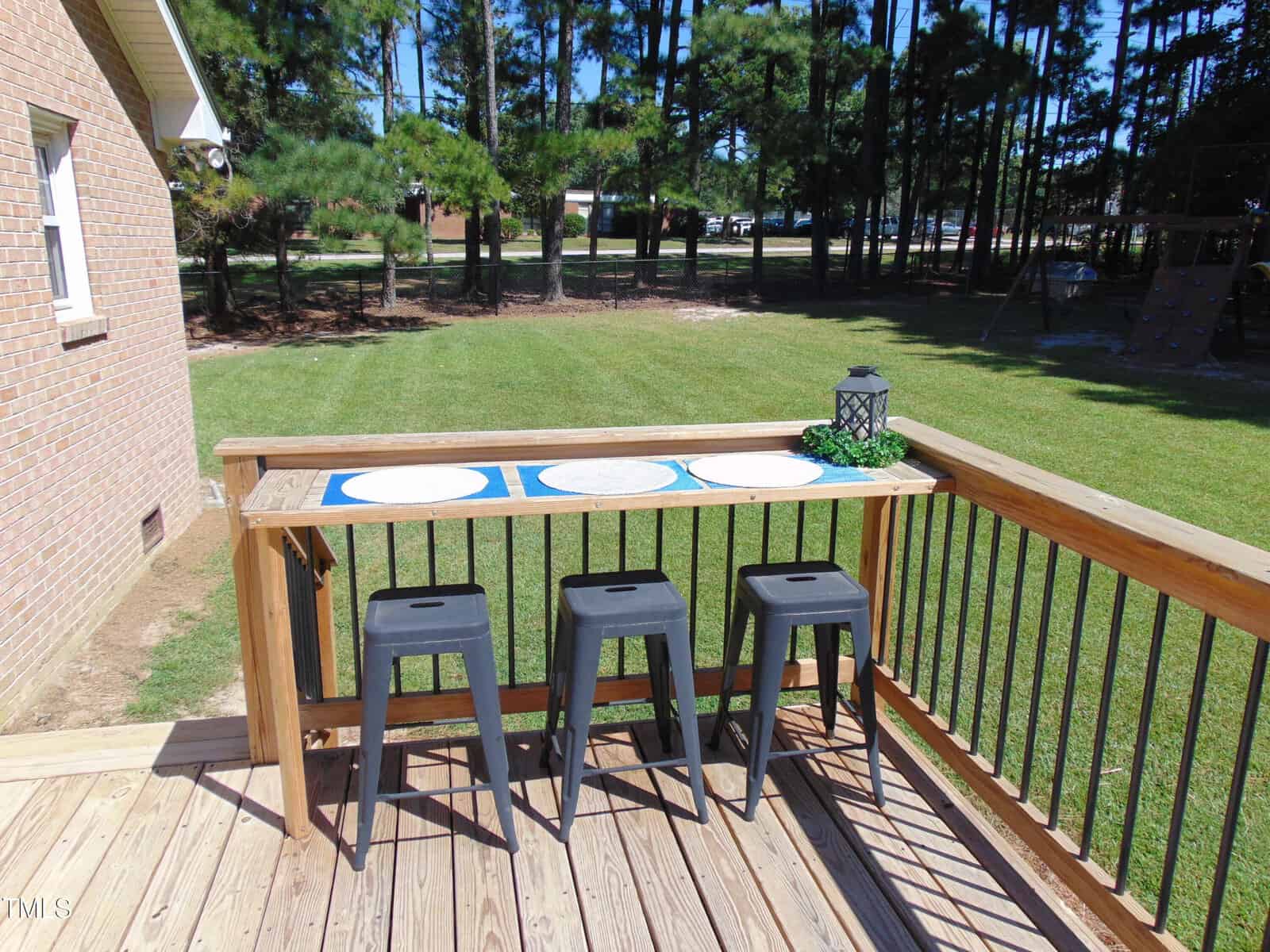 A wooden deck with a tall narrow table, three black stools, placemats, and a lantern centerpiece overlooks a grassy backyard bordered by tall pine trees.