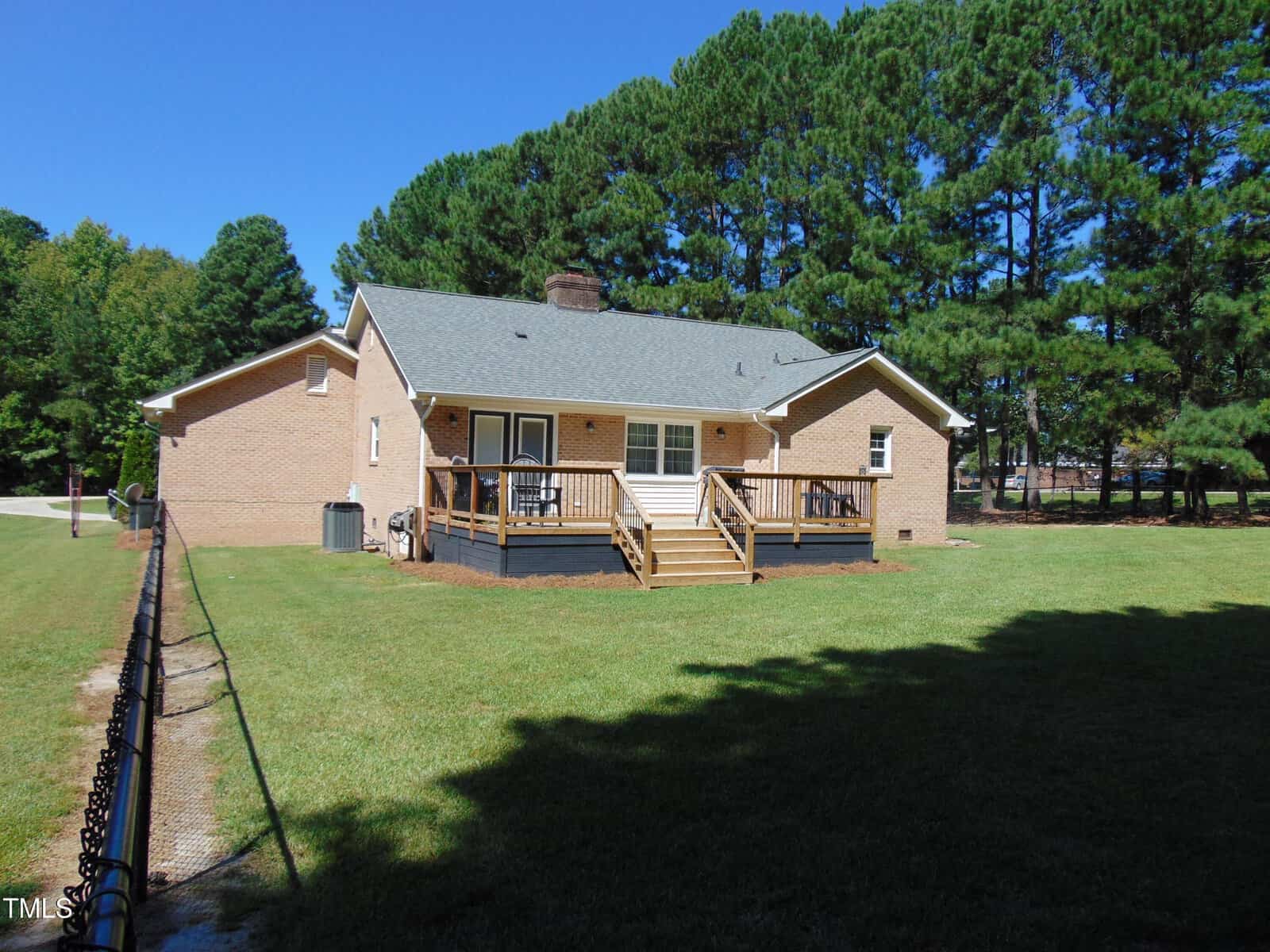 A brick house with a wooden deck and steps in the backyard, surrounded by a grassy lawn and a black chain-link fence, with tall pine trees in the background under a clear blue sky.