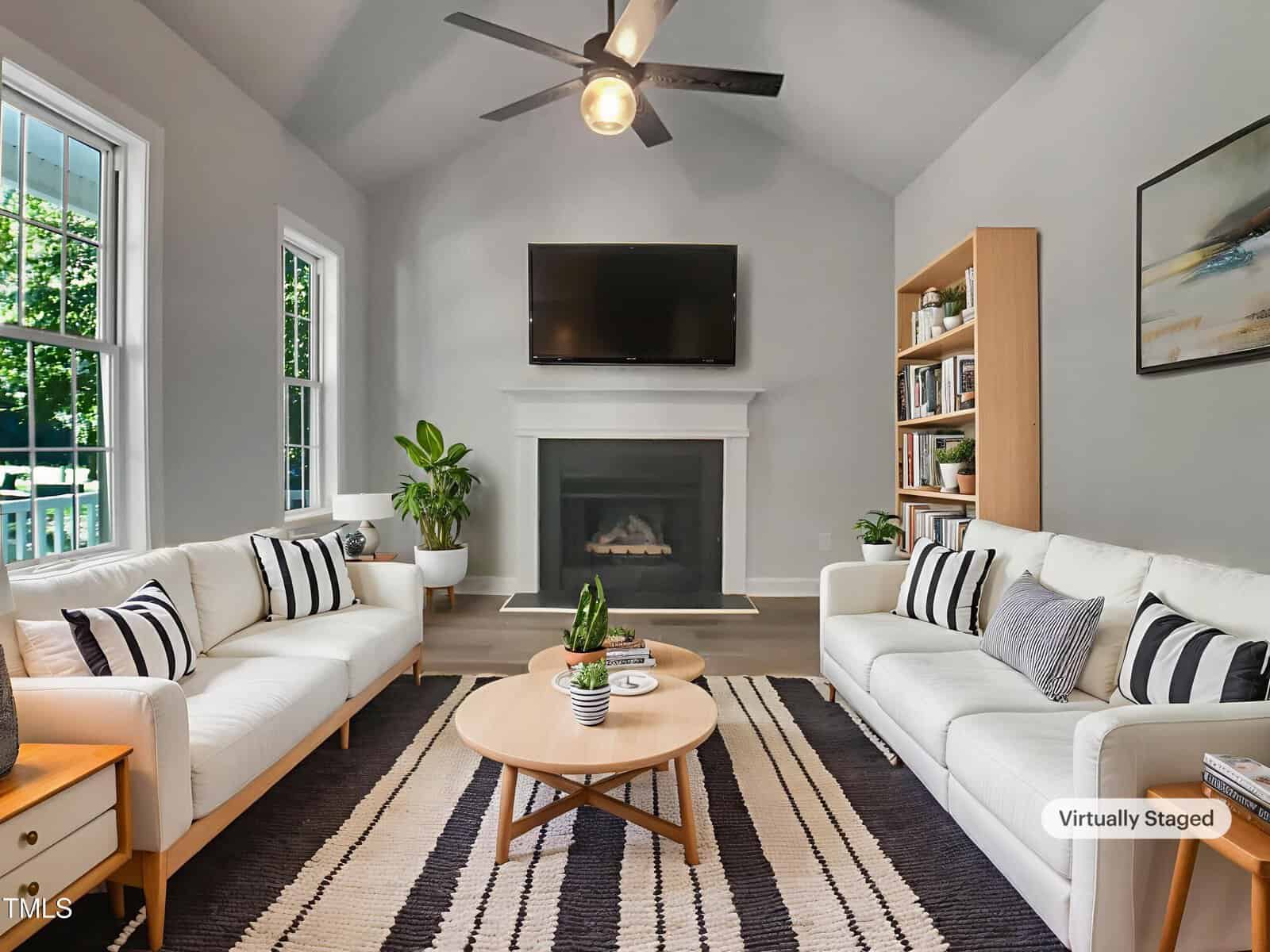 Modern living room with two white sofas, a wooden coffee table, a black-and-white rug, fireplace with mounted TV, tall bookshelf, potted plants, and gray walls. A ceiling fan hangs from the vaulted ceiling.