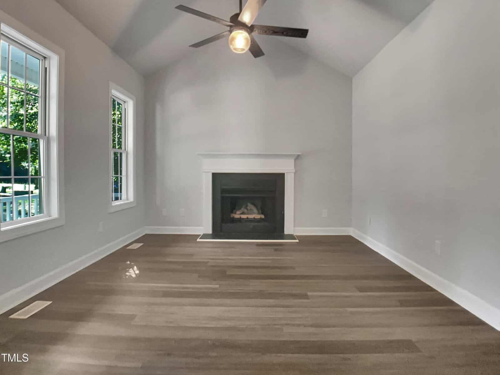 Empty room with gray walls, vaulted ceiling, a ceiling fan, and wood flooring. The room features a white-trimmed fireplace centered on the far wall and two windows on the left, showing greenery outside.