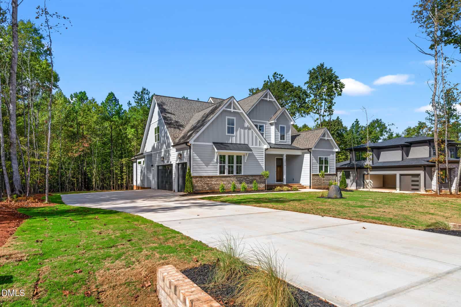 A modern two-story gray house with white trim, a large driveway, three-car garage, landscaped front yard, and surrounded by tall trees under a clear blue sky.