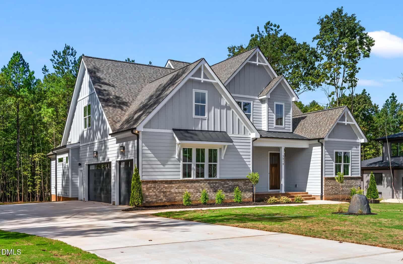 A modern two-story house with gray siding, white trim, gable roofs, and brick accents, featuring a double garage and a neatly landscaped yard, set against a backdrop of tall trees and clear blue sky.