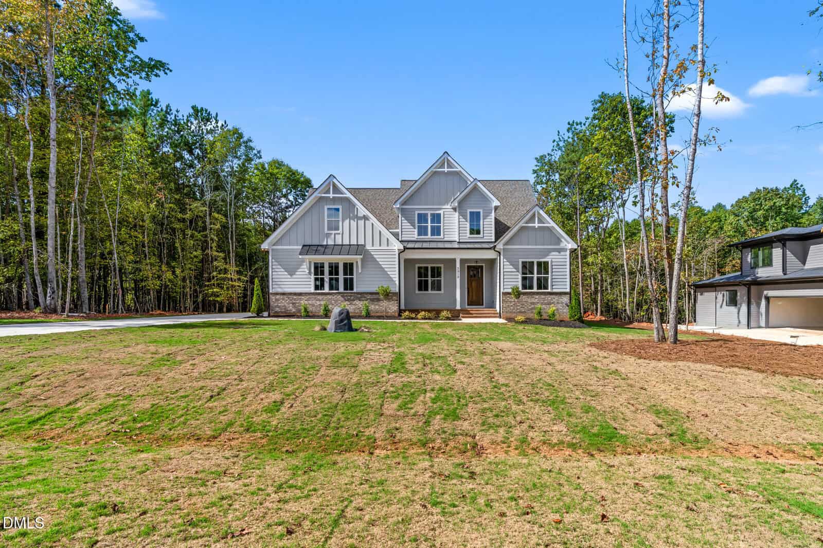 A modern two-story house with gray siding and white trim sits on a spacious, freshly sodded lawn, surrounded by tall trees under a clear blue sky. An attached garage is visible to the right.