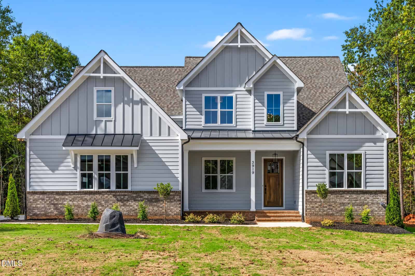 A modern two-story house at 3919 Cedar Knolls Drive in Youngsville, with gray siding, white trim, and a brick foundation, features multiple gables, large windows, and a wooden front door surrounded by green lawn and trees.