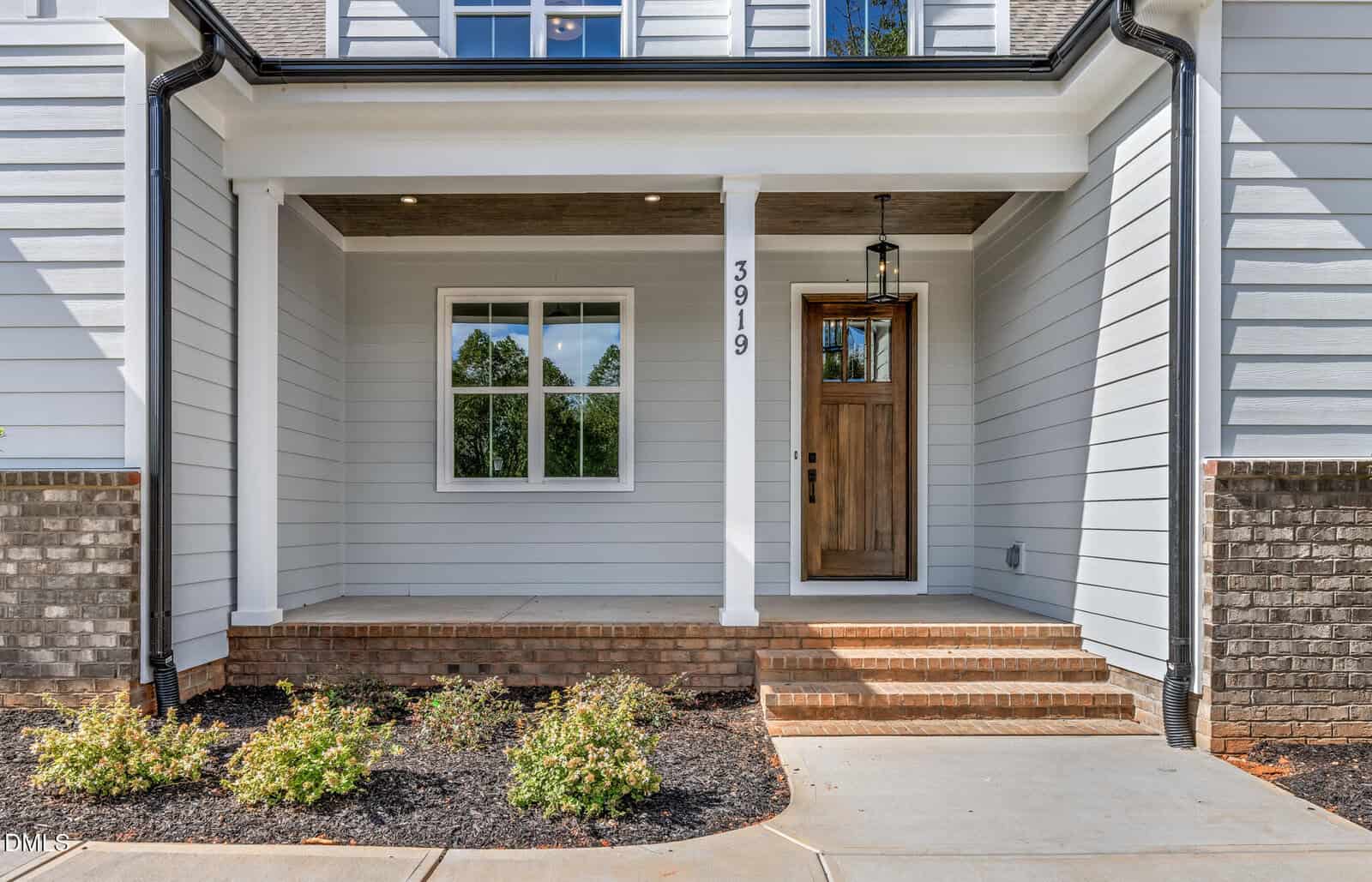 Front porch of a modern house with white siding, a wooden door with glass panel, a hanging lantern, brick steps, a column, and small shrubs planted along the walkway. House number 3919 is displayed vertically by the door.