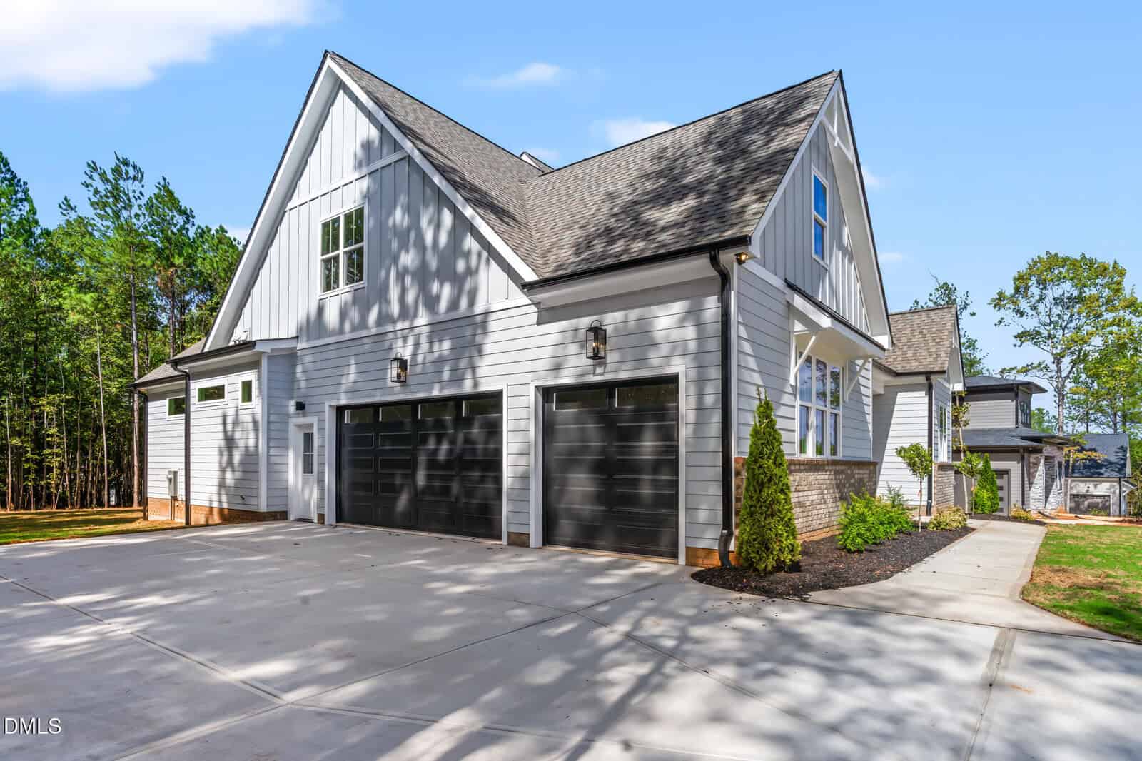 Modern white two-story house with three black garage doors, gable roof, multiple windows, and a large concrete driveway, surrounded by trees and landscaped greenery.