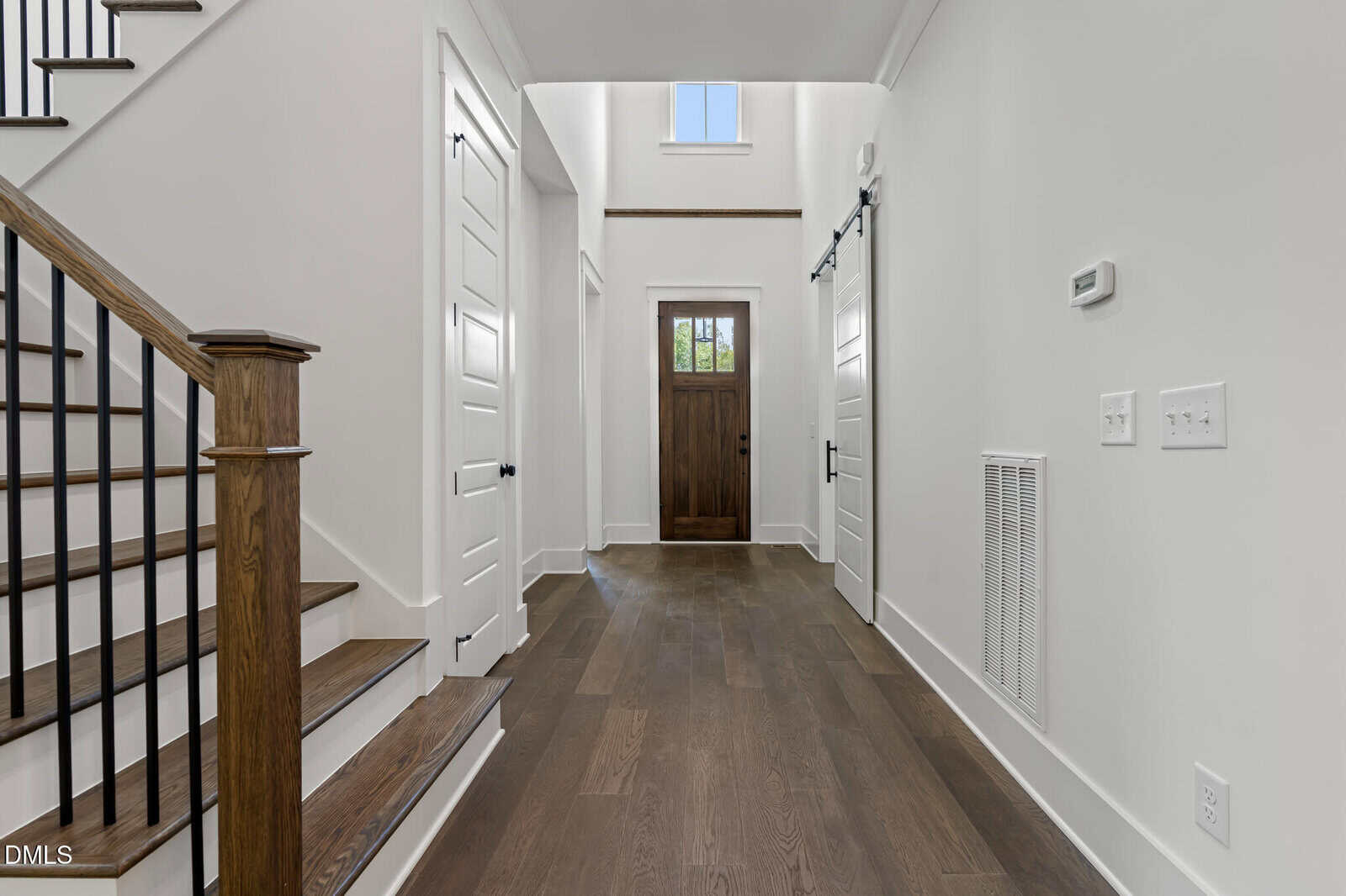 Bright hallway with wood flooring, white walls, and a wooden front door. Stairs with dark wood and black railings are on the left; several white doors and a small window are visible.