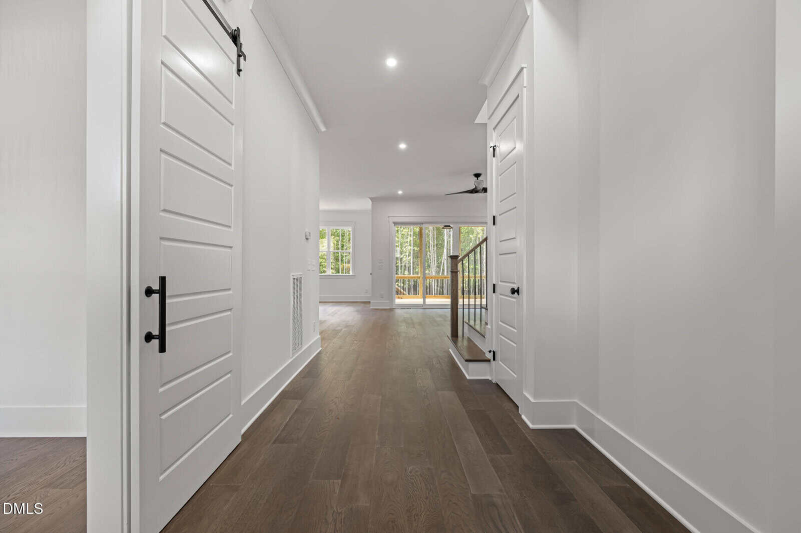Bright hallway with white walls and doors, dark wood floors, leading to an open living area with large windows and a view of trees outside. Stairs with a railing are visible on the right.