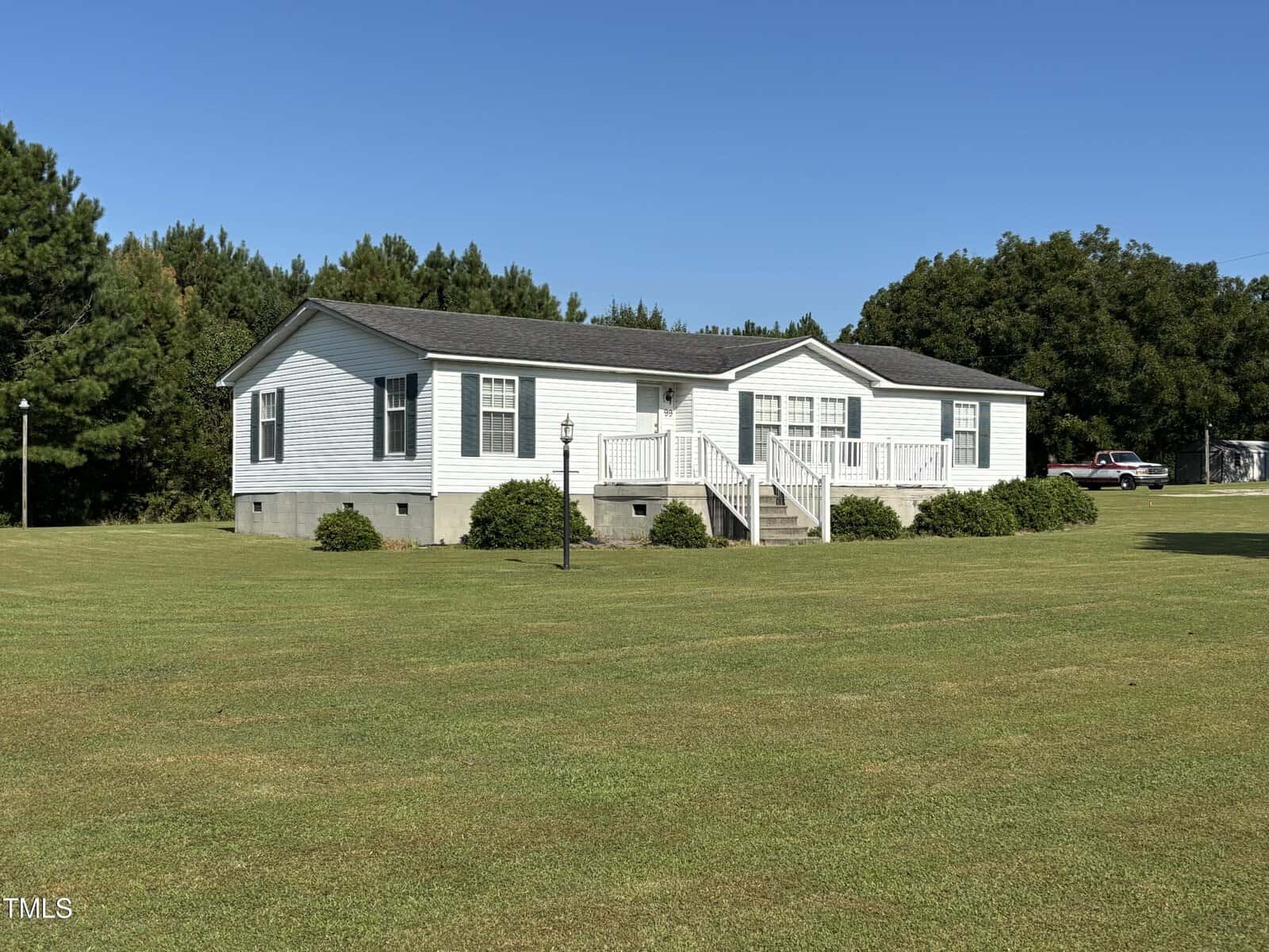 A white single-story manufactured home at 99 Kelly Road, Henderson features a front porch and steps, set on a large, well-manicured lawn with bushes along the foundation and surrounded by trees under a clear blue sky.