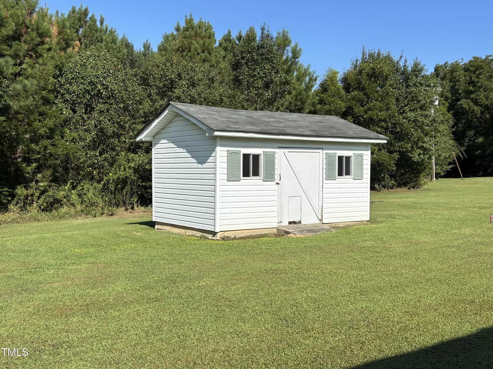 A small white shed with gray roof, green shutters, and a double door sits on a grassy lawn with trees and bushes in the background under a clear blue sky.