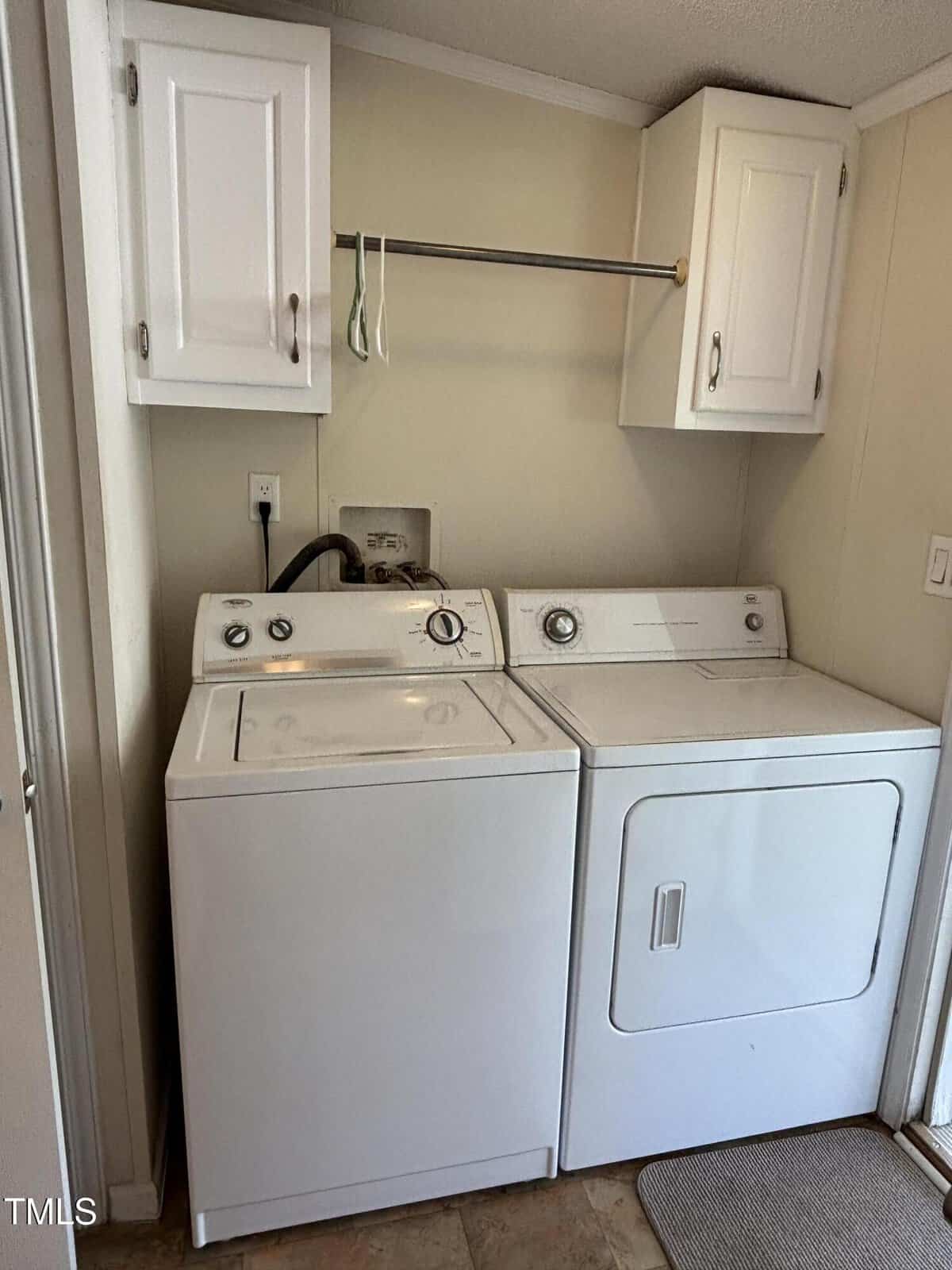 A laundry area with a white washing machine and dryer side by side, white cabinets above, a metal hanging rod, and beige walls. There is a wall outlet and hoses connected behind the machines.