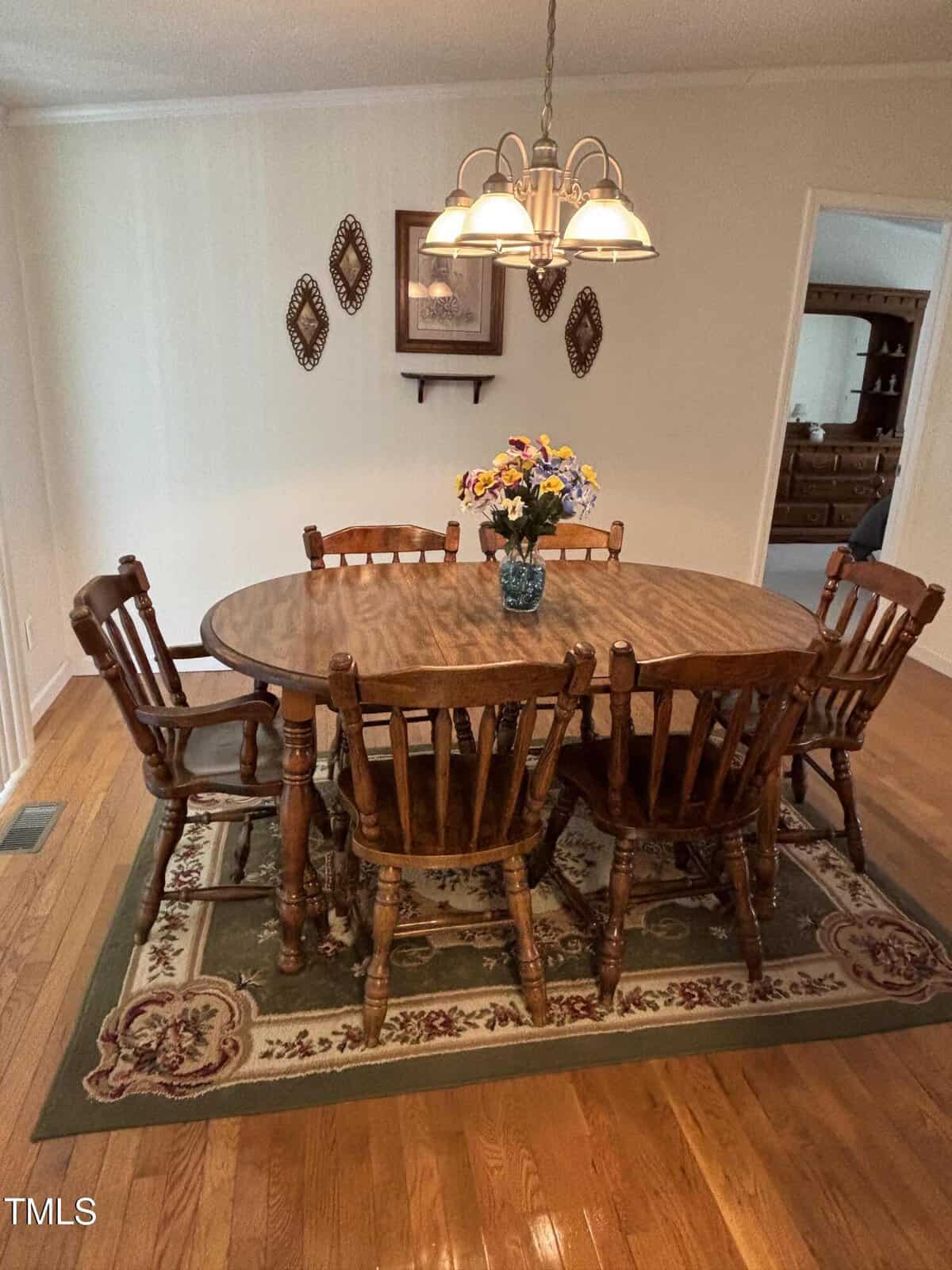 A wooden dining table with six matching chairs sits on a patterned rug. A vase with colorful flowers is centered on the table. Decorative wall art and a light fixture are above. Hardwood floor and neutral walls surround the area.