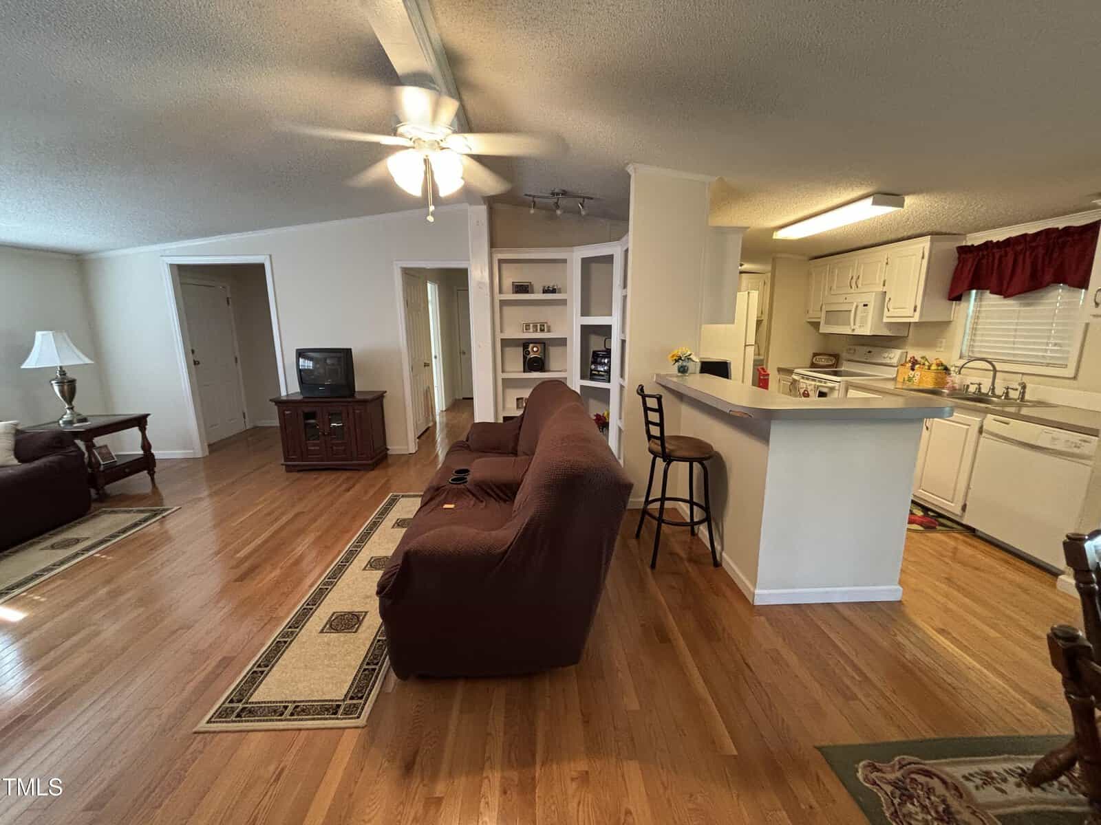 A living room with hardwood floors, brown couches, a TV on a wooden stand, and a kitchen with white cabinets and appliances. A breakfast bar with two stools separates the kitchen from the living area.