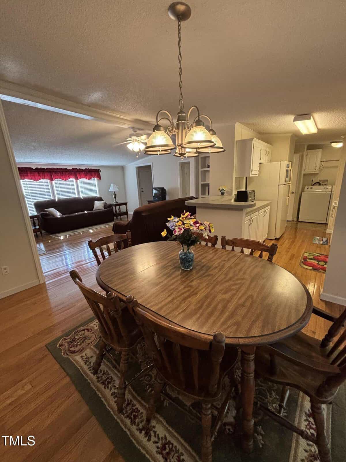 A dining area with a wooden table and chairs sits on a rug, next to a kitchen with white cabinets. In the background is a living room with a brown sofa, armchair, coffee table, and large window with red curtains.