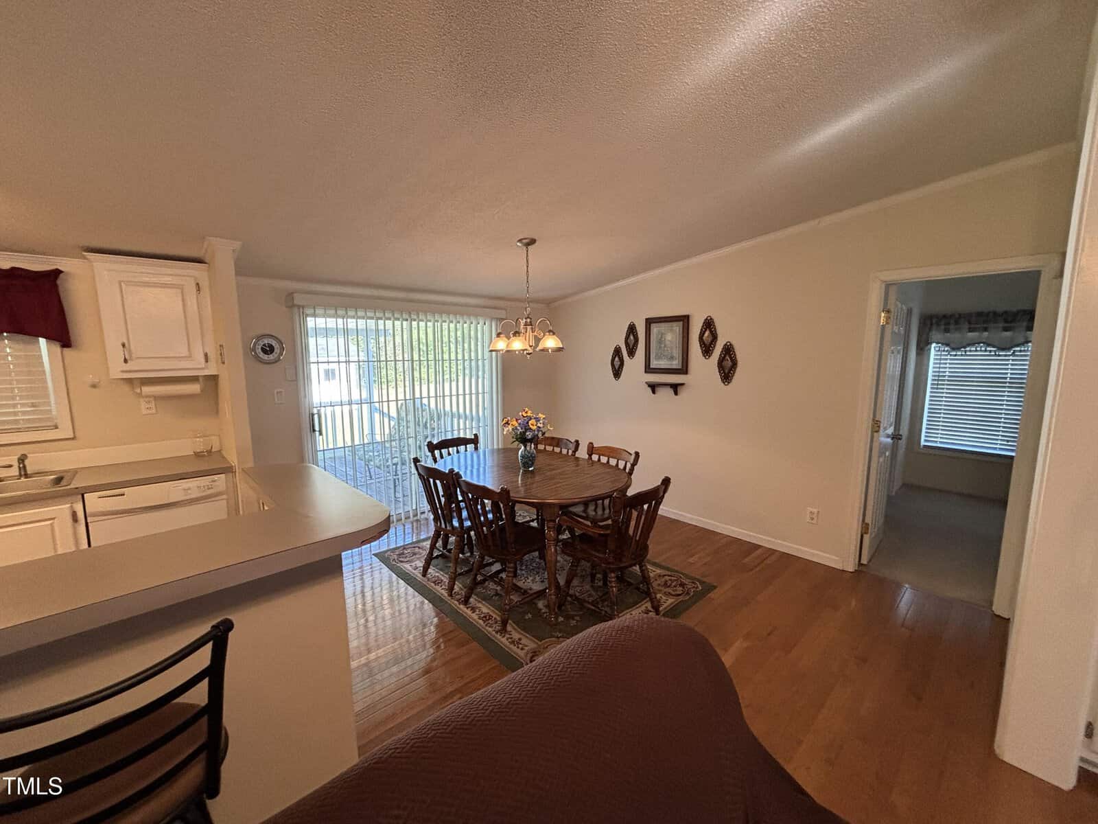 A dining area with a wooden table and six chairs is set near a sliding glass door leading outside. A chandelier hangs above the table. To the left is a partial view of a kitchen, and a doorway leads to another room.