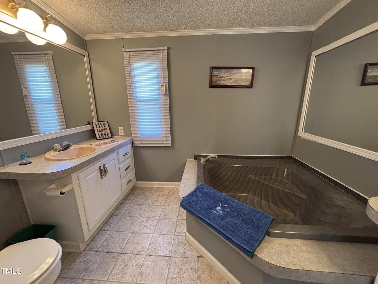 Bathroom with a large black jacuzzi tub, gray walls, a white vanity, mirror, window with blinds, and a blue towel on the tub ledge. Framed art and sign reading “Live, Laugh, Love” are visible.