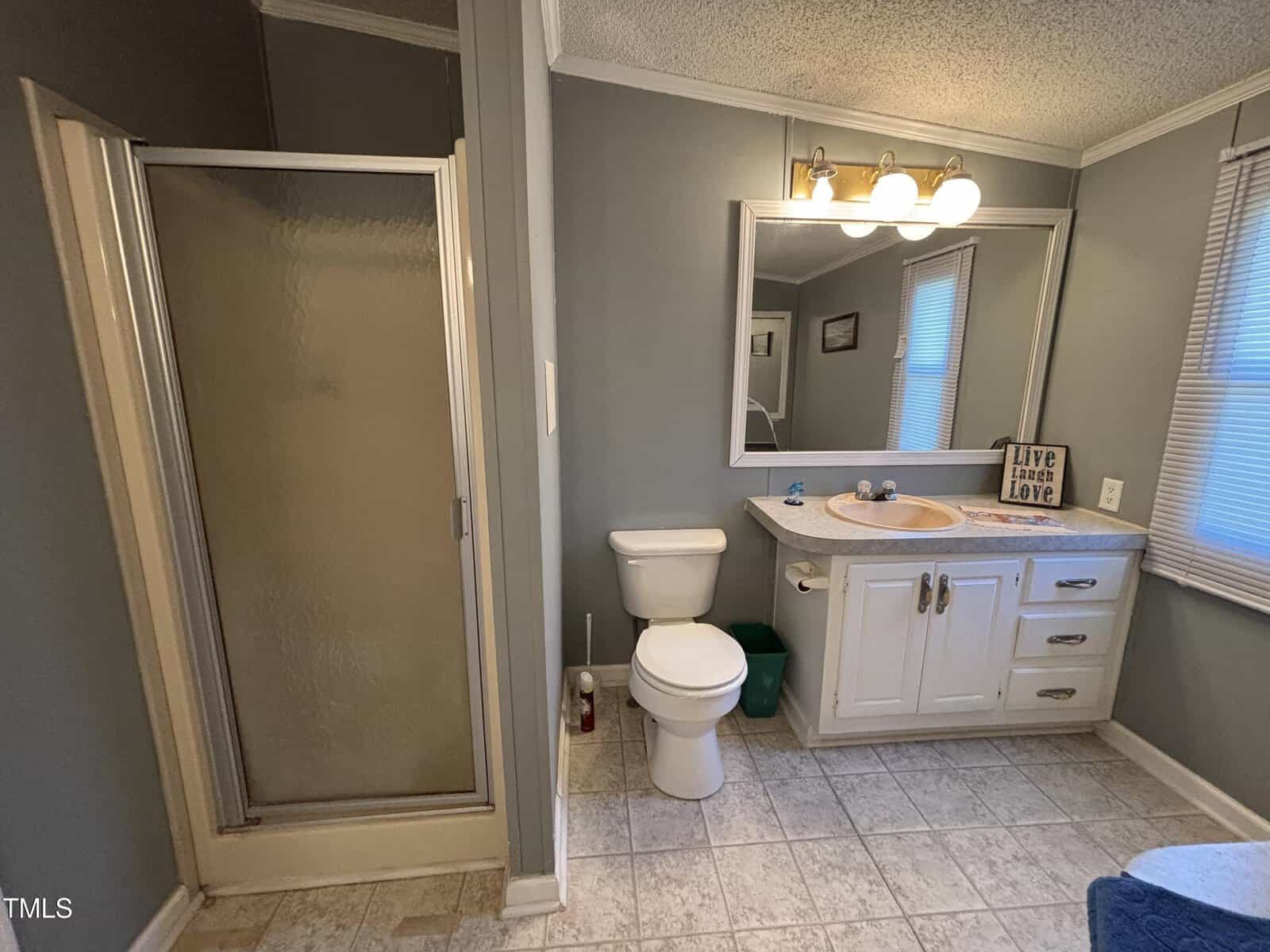 A bathroom with a shower, toilet, sink, white vanity, large mirror, three-light fixture, and a window. The floor is tiled, and the walls are painted gray. A sign reading “Live, Laugh, Love” sits on the counter.