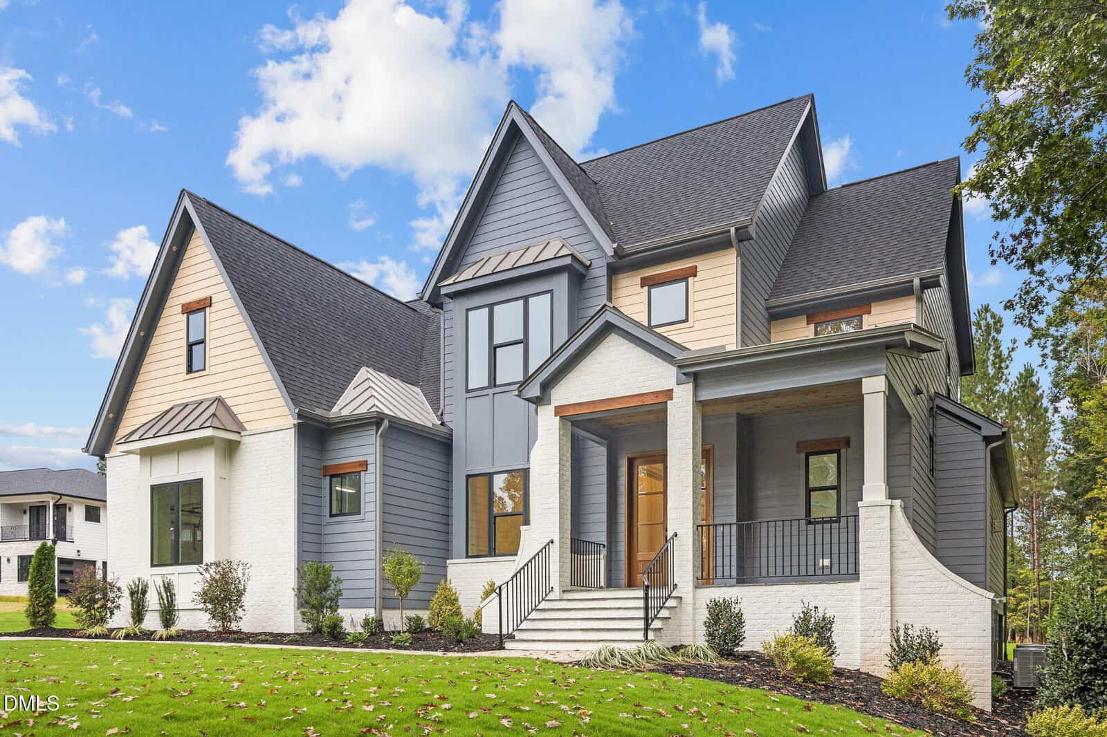Modern two-story house with gray and beige siding, white brick accents, a covered front porch, large windows, and neatly landscaped front yard under a blue sky with scattered clouds.