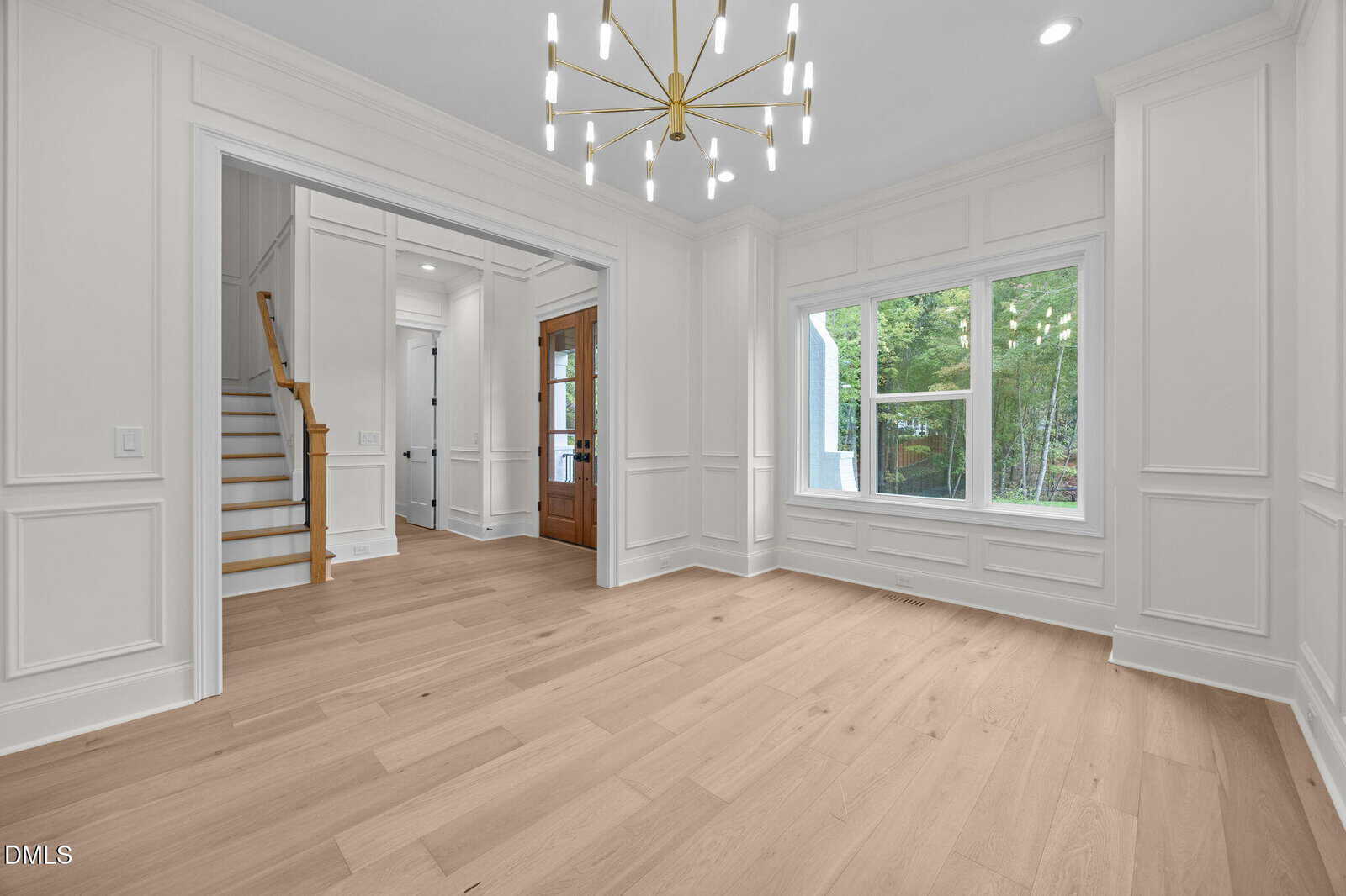 A bright, modern entryway with light wood floors, white paneled walls, large windows, and a gold chandelier. A wooden front door and staircase are visible in the background.
