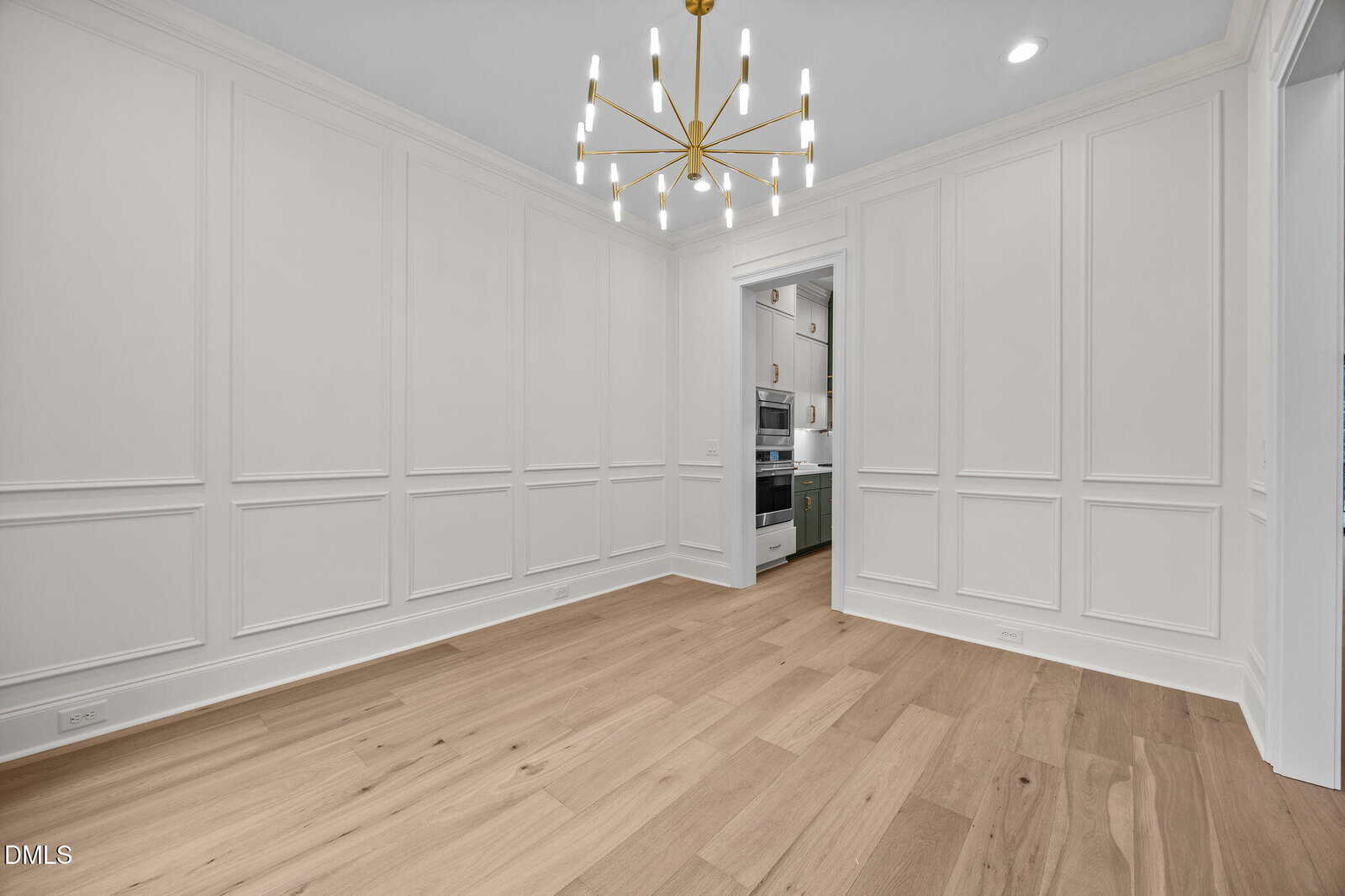 A modern, empty dining room with white paneled walls, light wood flooring, and a gold chandelier. An open doorway leads to a kitchen with white cabinetry and built-in appliances.