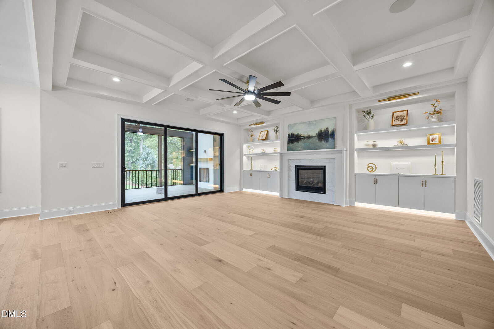 Bright, modern living room with light wood floors, coffered ceiling, built-in shelves around a fireplace, and large sliding doors opening to a covered porch with views of trees outside.