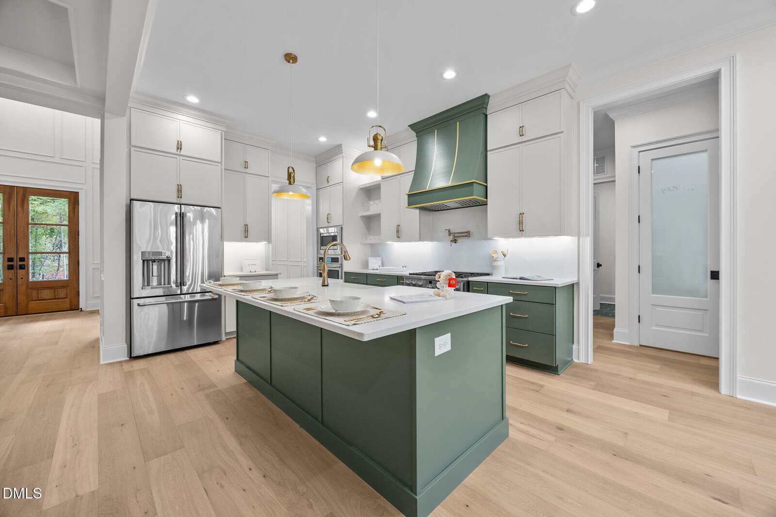 A modern kitchen with green and white cabinetry, a green island with bar stools, stainless steel appliances, pendant lights, and light wood flooring. There's a double-door fridge and a door to a pantry on the right.