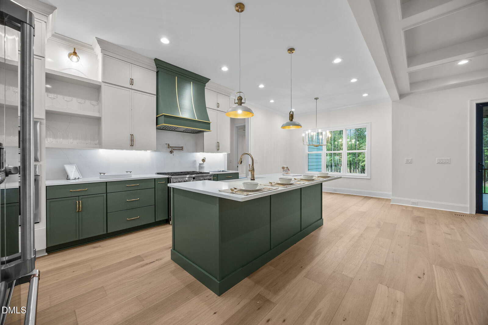 Modern kitchen with green cabinets and island, white countertops, gold fixtures, pendant lights, light wood floors, and large windows letting in natural light.
