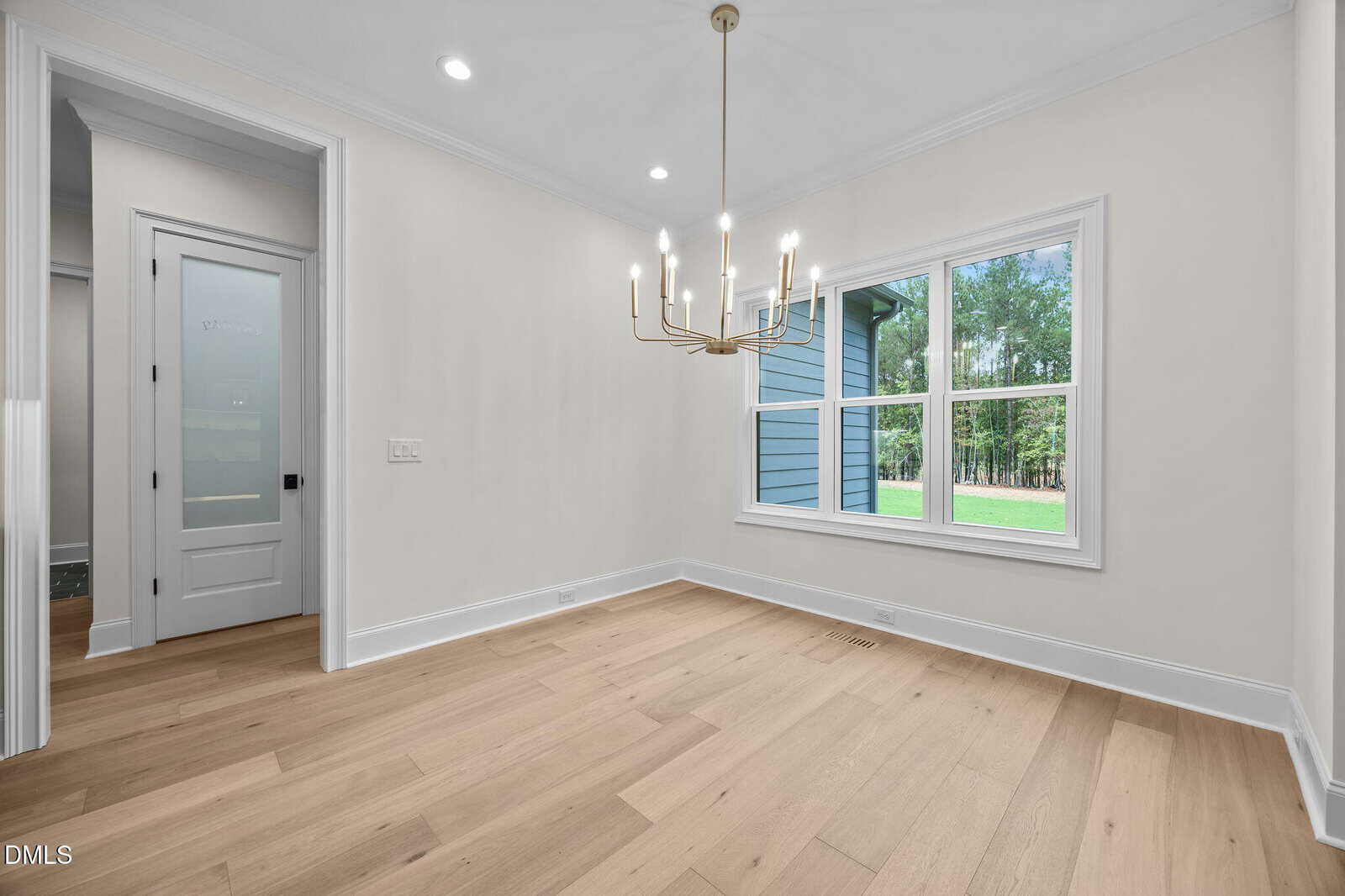 A bright, empty room with light wood flooring, white walls, a modern gold chandelier, a large window showing trees and grass outside, and a door with frosted glass on the left.