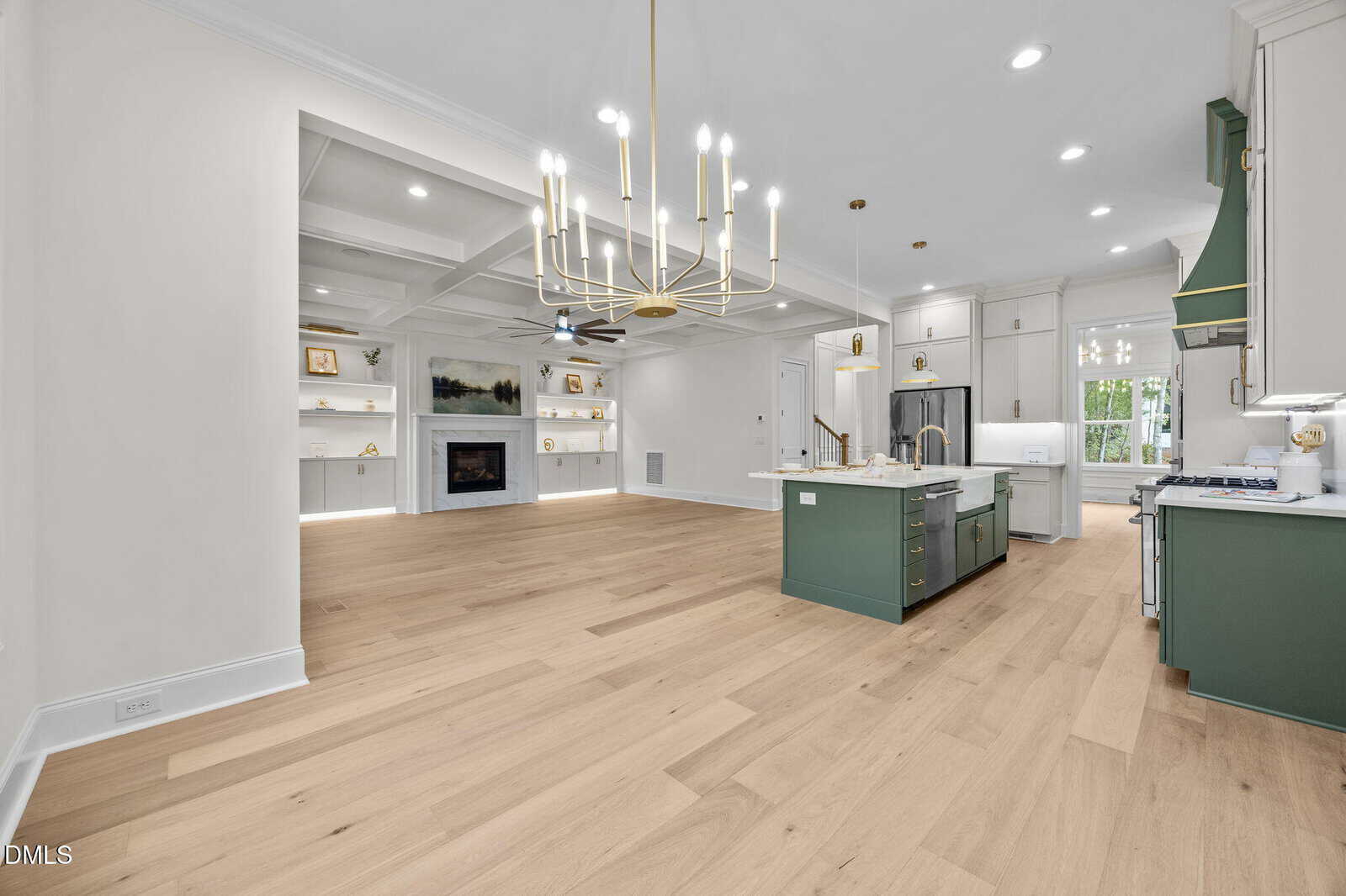Modern kitchen with light wood floors, green island, white cabinets, gold chandelier, stainless appliances, and open living space with built-in shelves, a fireplace, and large windows.