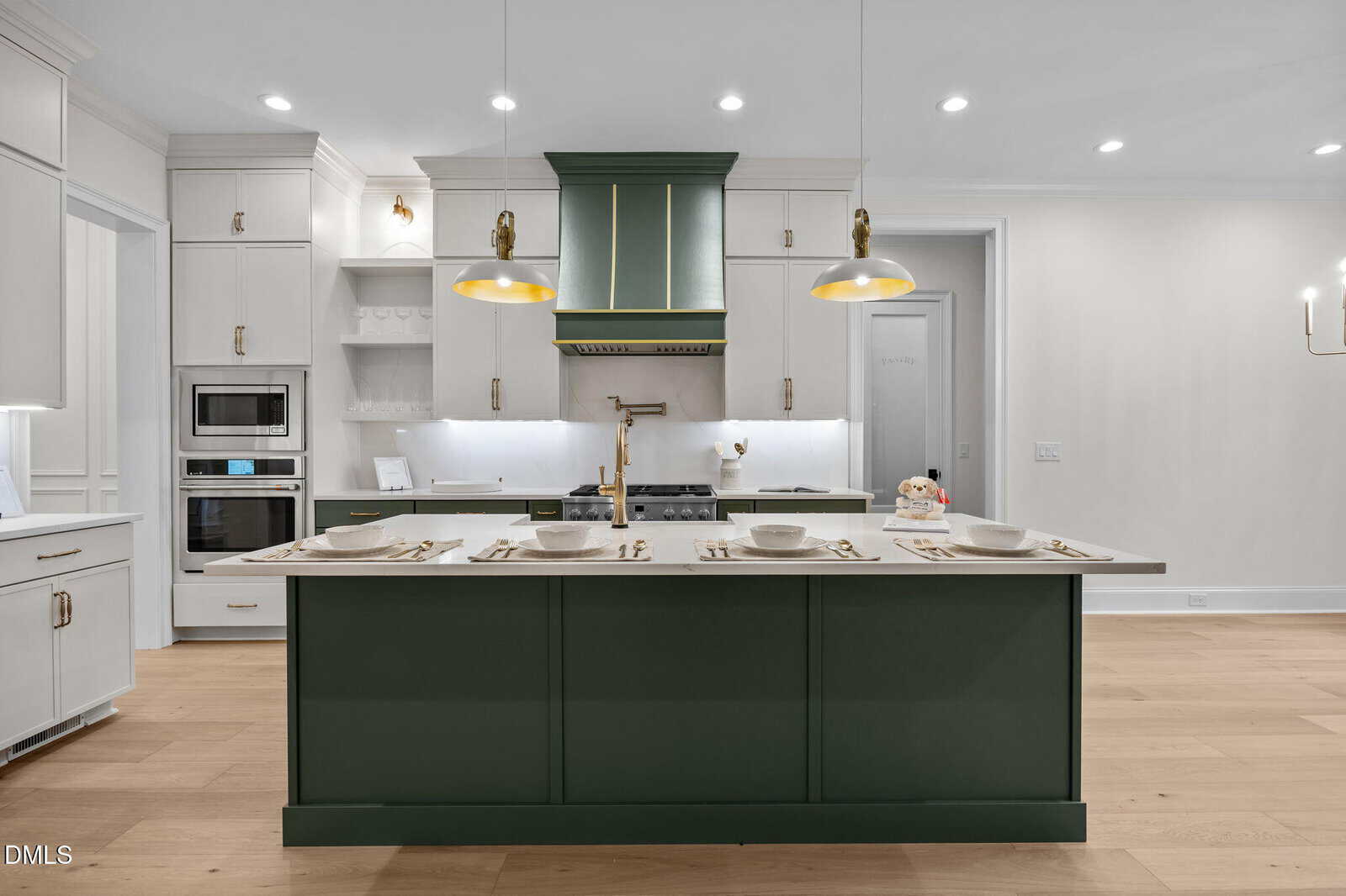 Modern kitchen with a large green island, three place settings, gold and white pendant lights, white cabinets, built-in oven and microwave, light wood flooring, and a sleek vent hood above the stove.