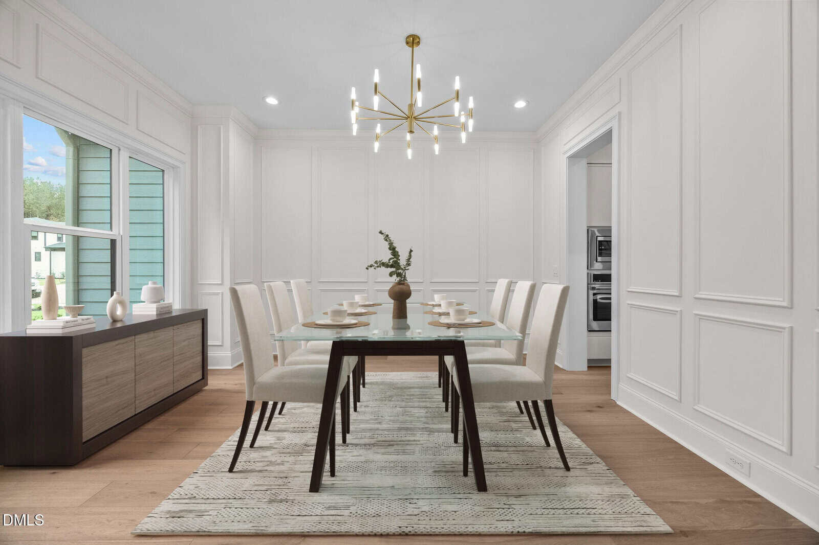 Modern dining room with a glass-top table, eight cream chairs, a geometric gold chandelier, sideboard with decor, large window, and neutral walls with molding. A vase with greenery sits on the table. Light wood flooring throughout.