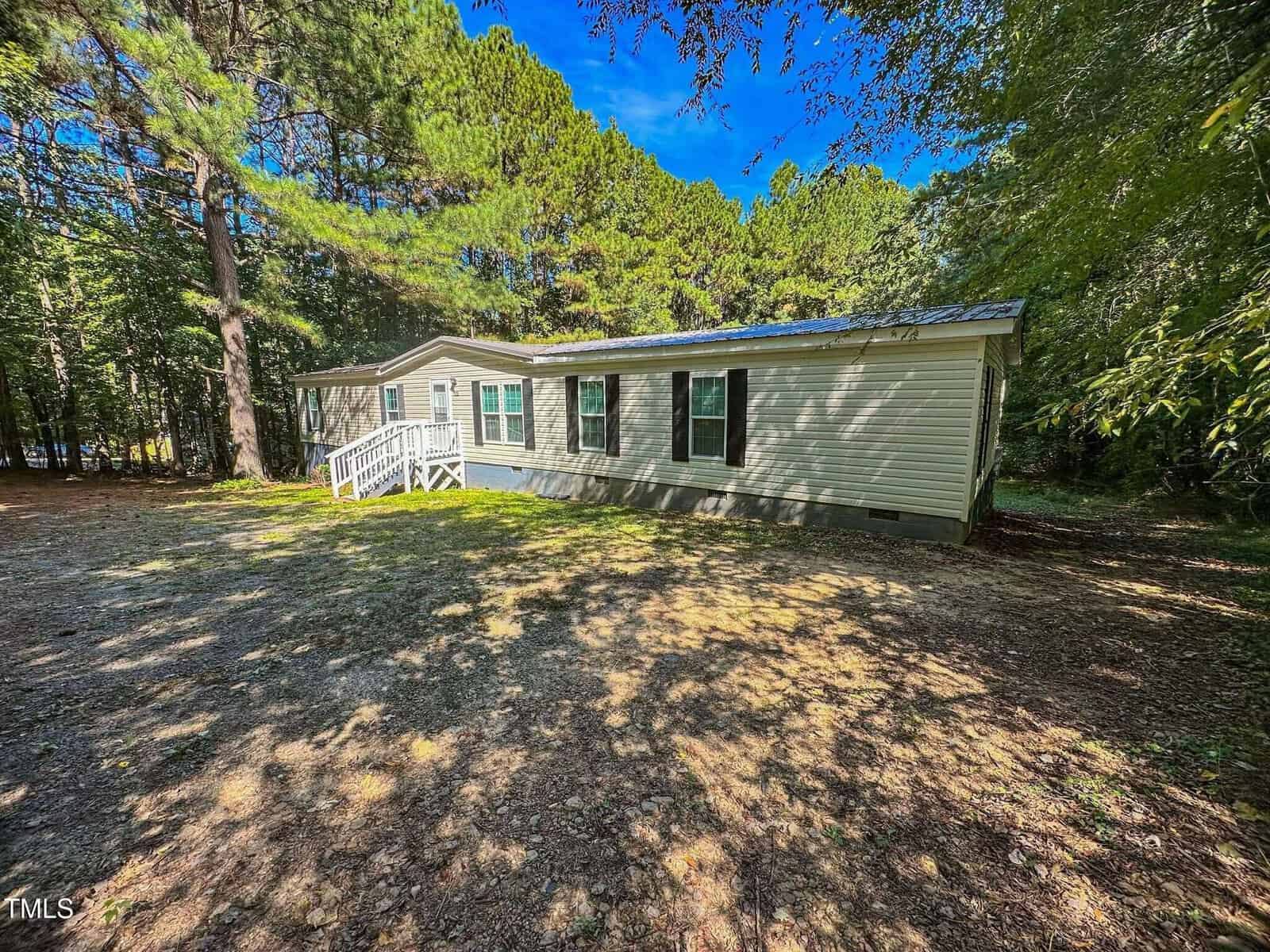 A single-story manufactured home with light-colored siding, black shutters, and a small porch sits on a shaded lot surrounded by tall trees. Sunlight filters through the trees onto the yard and house.