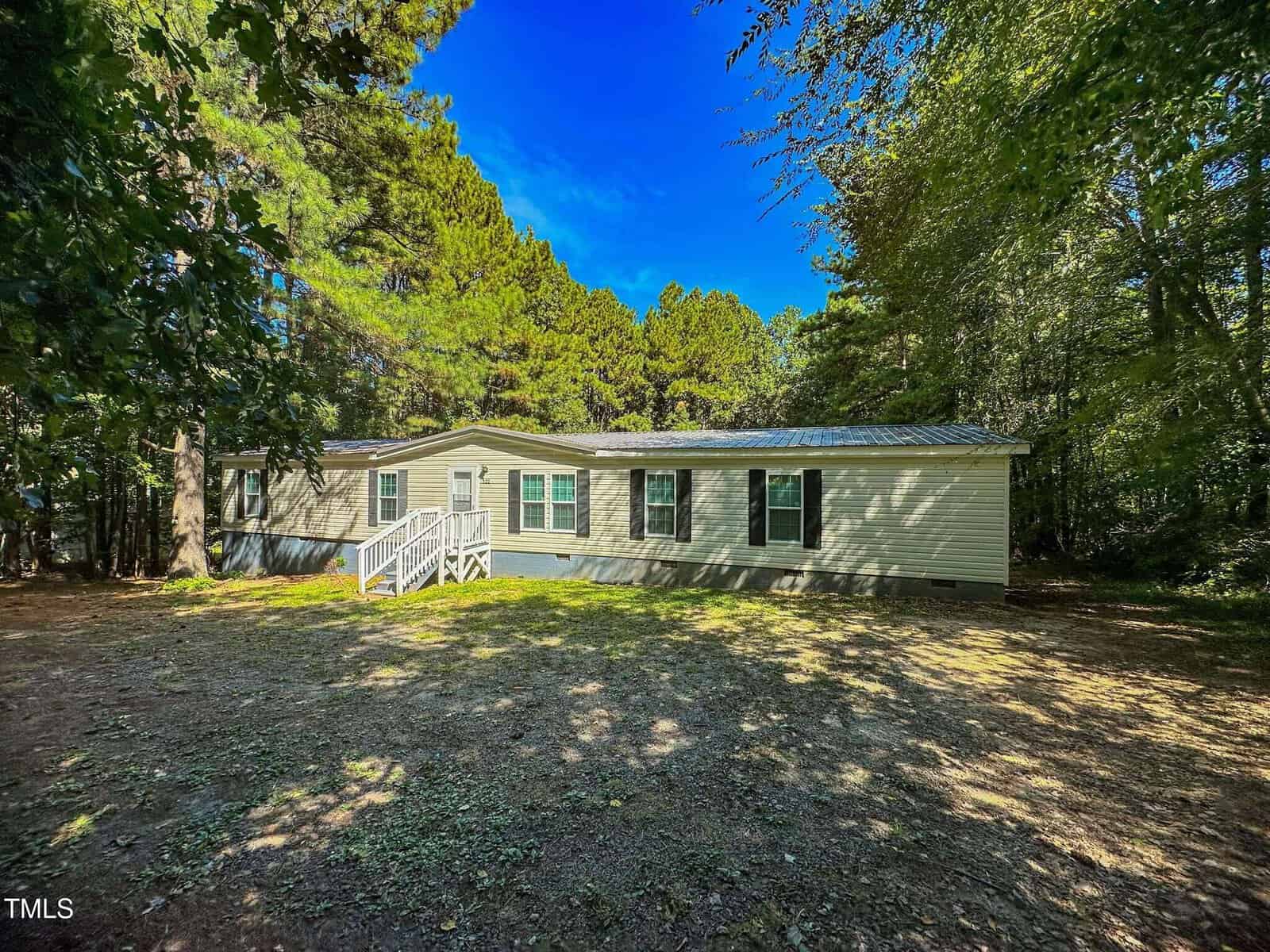 A single-story manufactured home with beige siding and green shutters sits on a spacious, shaded lot surrounded by tall trees. A small white-railed porch with steps leads to the front door.