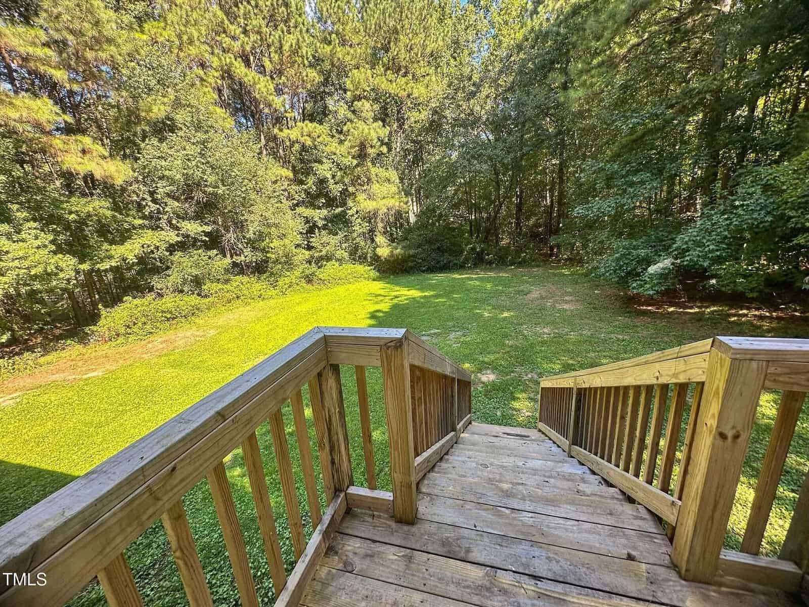 View from the top of a wooden staircase leading down to a large, grassy backyard surrounded by dense trees and greenery. Sunlight casts shadows on the lawn and trees.