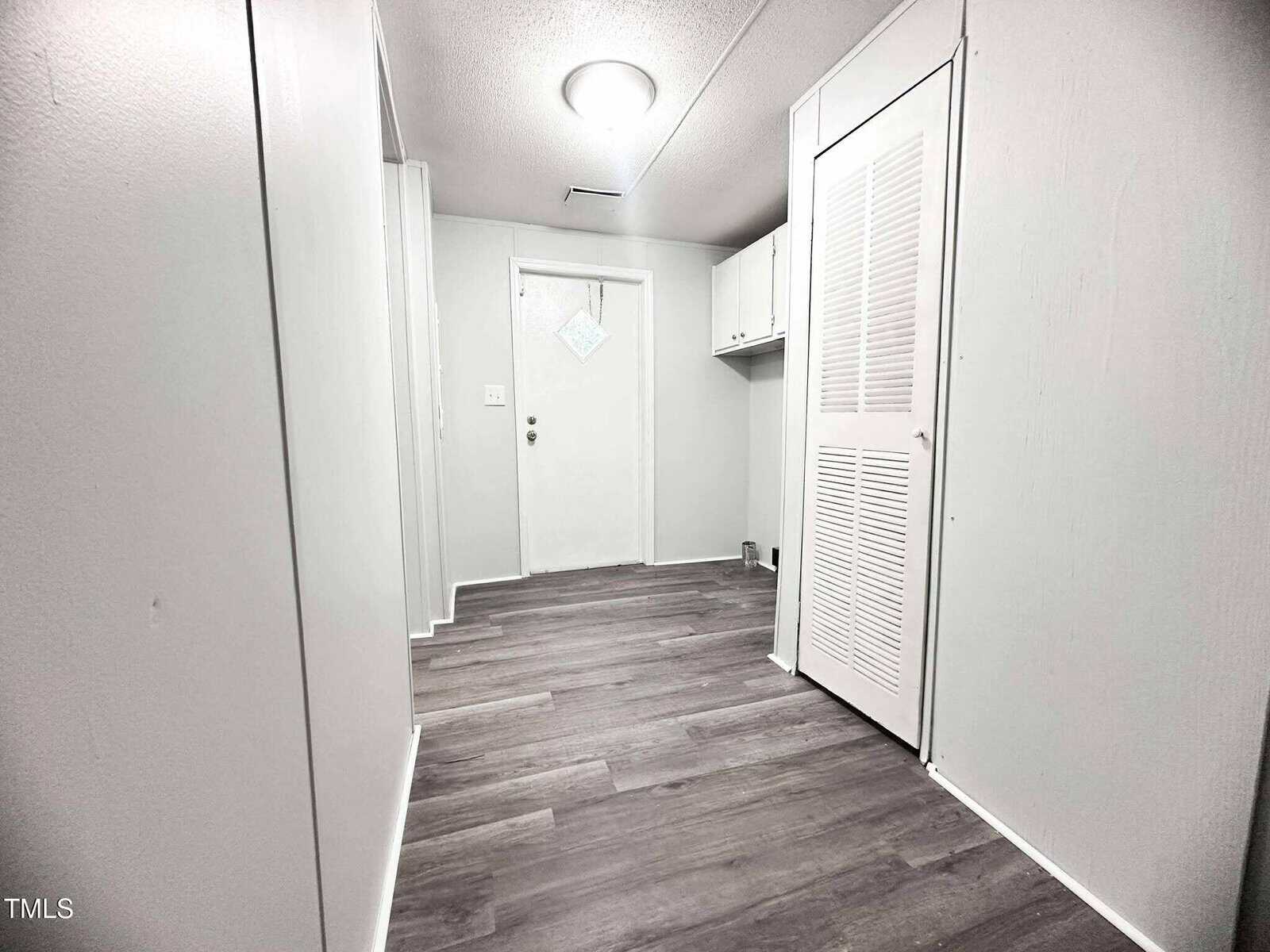 A hallway with light-colored walls and dark wood-style flooring leads to a white door. There is a louvered closet door on the right and overhead cabinets next to the door. A ceiling light fixture is centrally located.