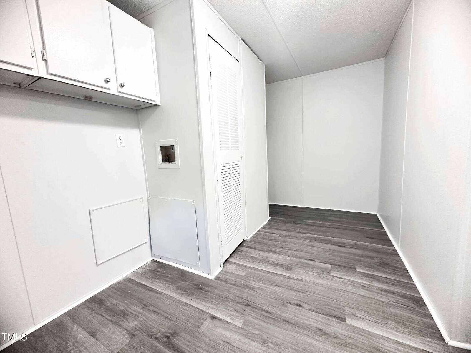 A small, empty room with light gray walls, white cabinets and doors, and dark gray wood-look flooring. There is a wall vent and an electrical outlet near the floor.