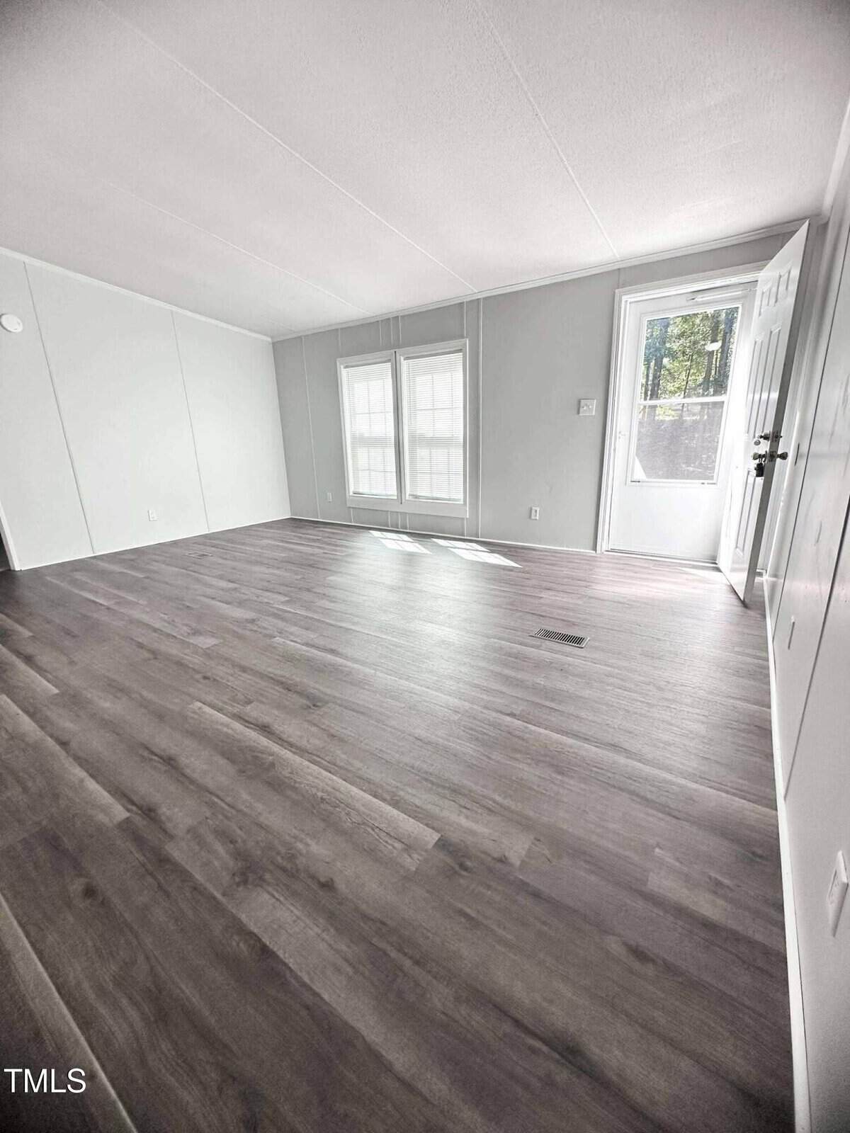 A bright, empty room with light gray walls, wide windows, and wood-style flooring. Sunlight streams through the windows, and an open door reveals an outside view.