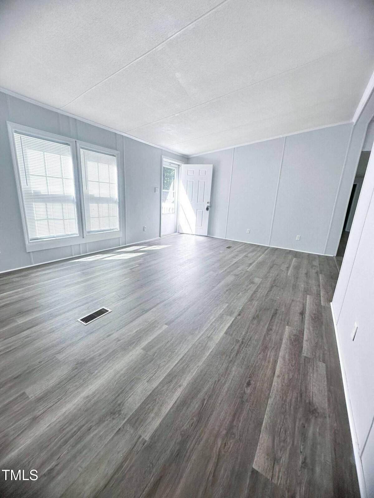 A bright, empty room with light gray walls, large windows, wood-look flooring, and a white front door. Sunlight streams in, giving the space an open, airy feel.