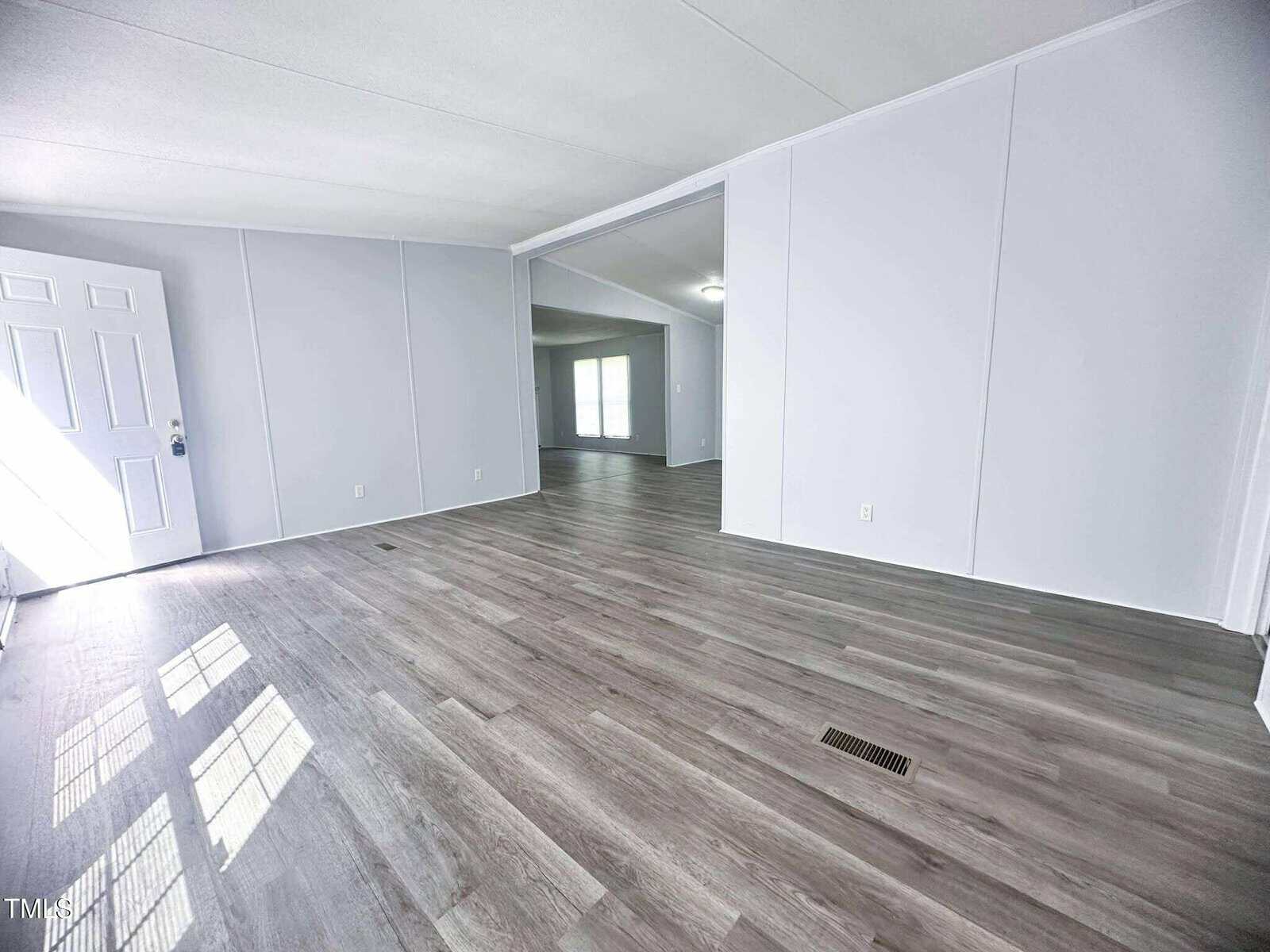 Bright, empty room with light gray walls, large windows, and wood-look vinyl flooring. An open doorway leads to another spacious area with similar flooring and lighting. Front door and vent visible.