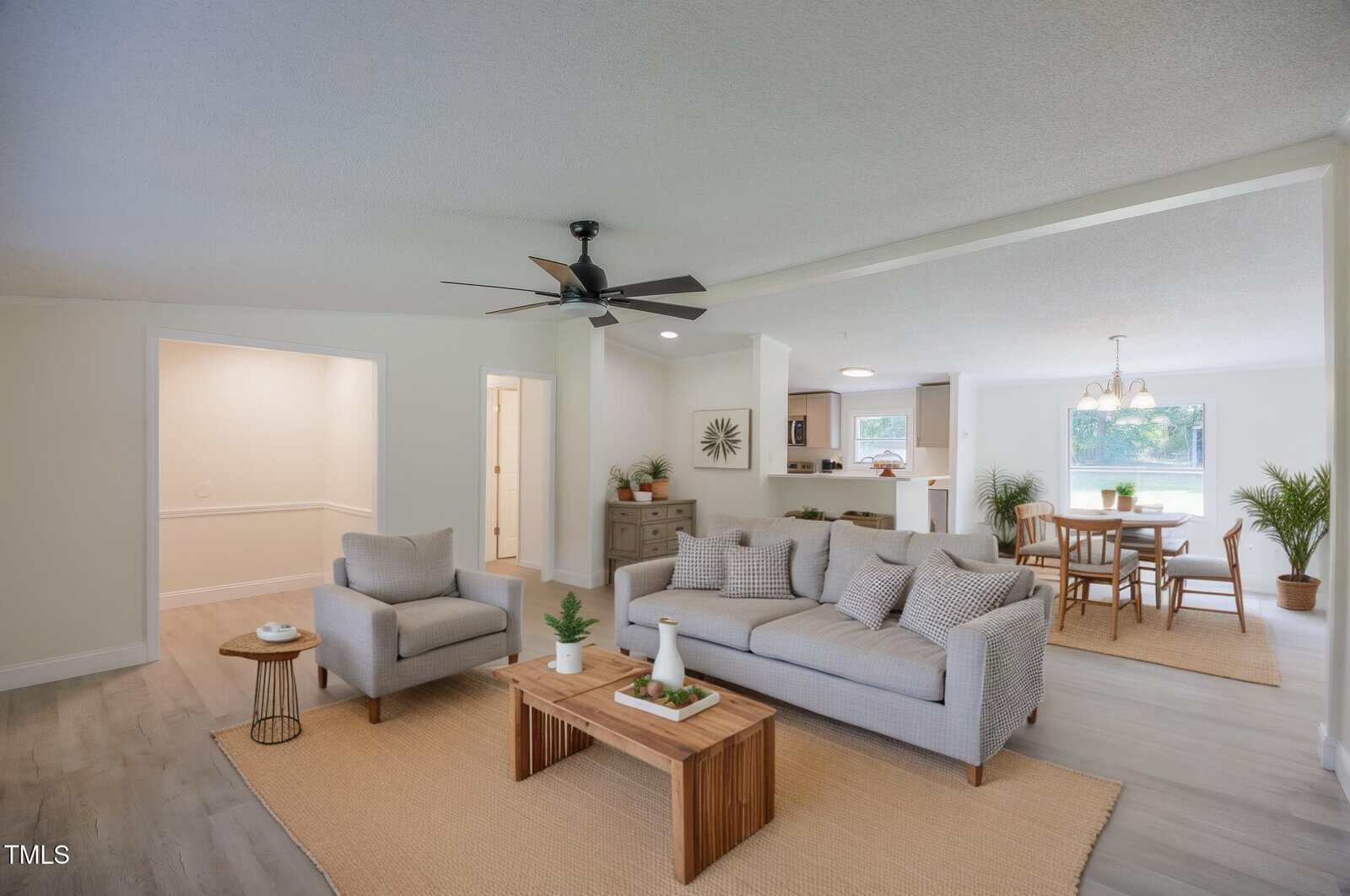 A bright, open living and dining area with neutral decor. There is a sofa, armchair, and wooden coffee table on a rug. The dining table and chairs are near large windows and indoor plants. A ceiling fan is above the seating area.