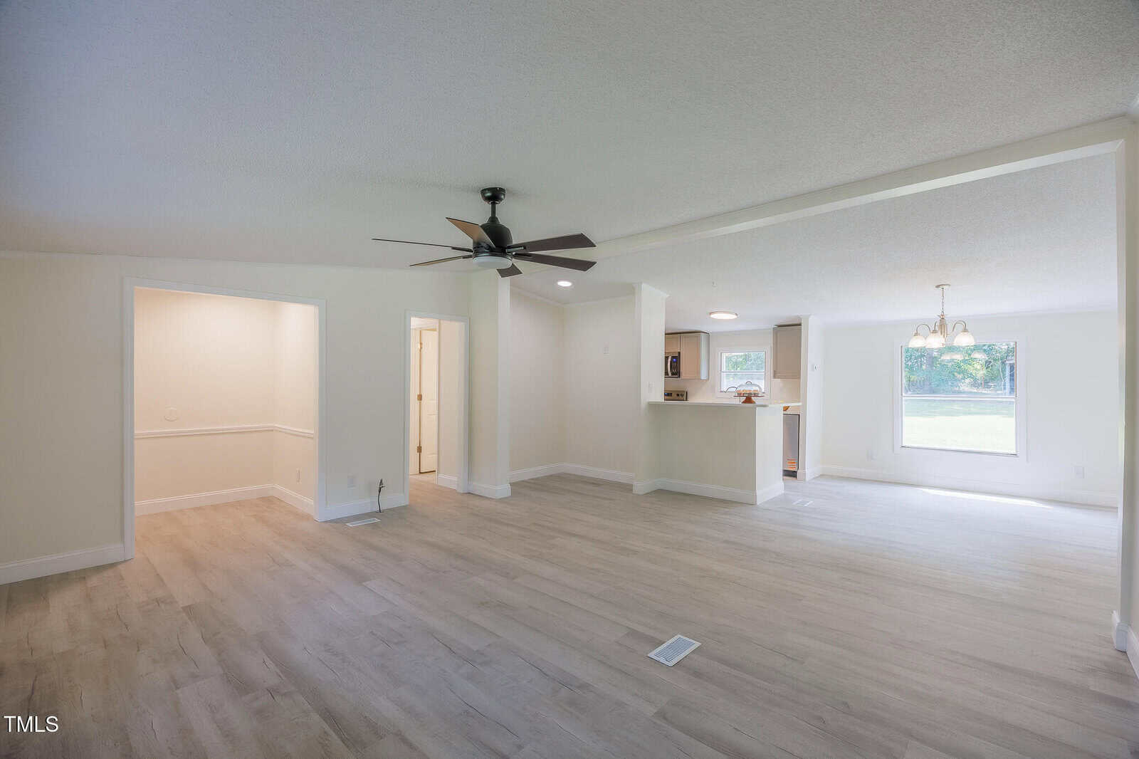 Spacious open-concept living area with light wood floors, ceiling fan, and adjacent dining area. Kitchen with breakfast bar visible; large window in dining space. Walls and trim are painted white.