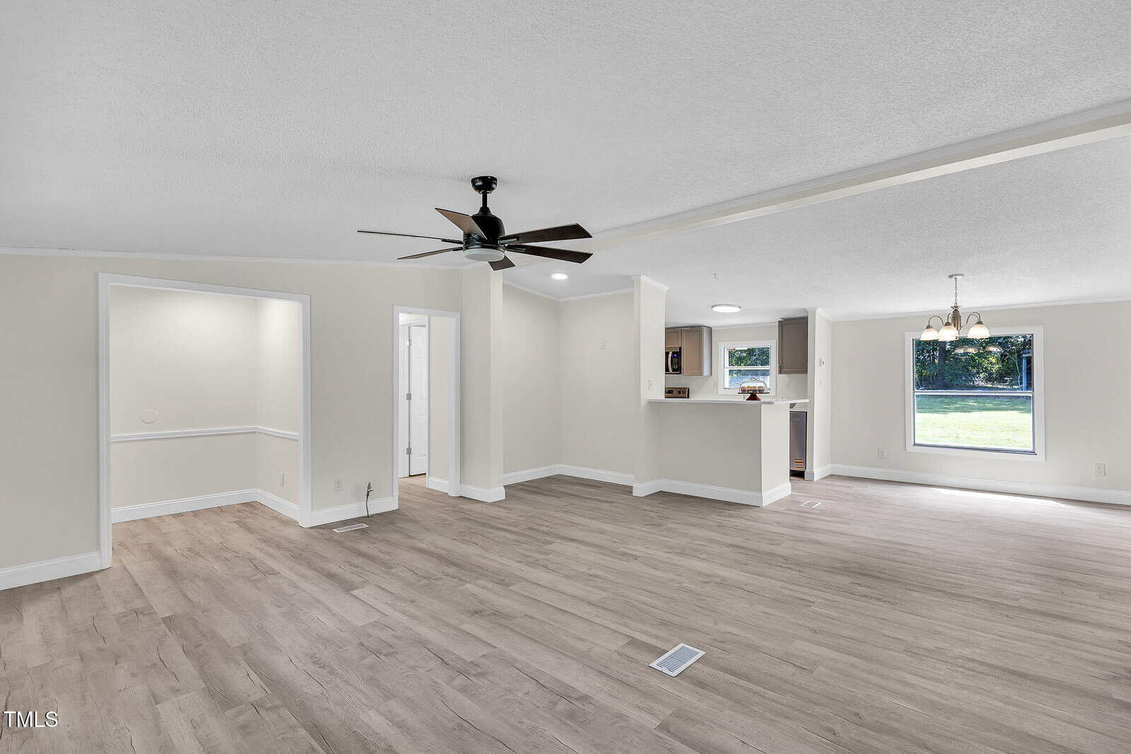 Spacious open living area with light wood flooring, neutral walls, ceiling fan, and large windows. Kitchen with pass-through opening and adjacent dining area visible in the background. Two doorways lead to other rooms.